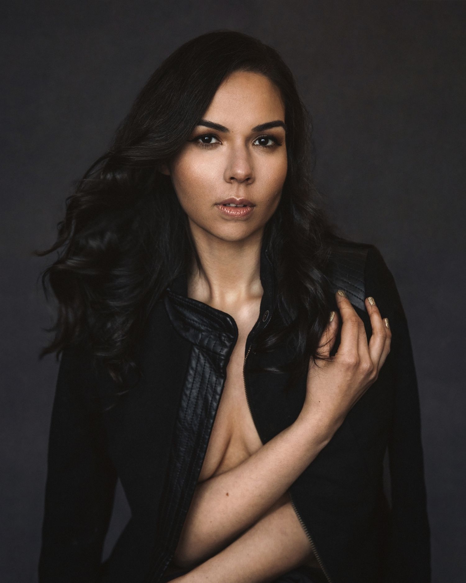 Dramatic portrait of a woman with flowing dark hair in black jacket against dark backdrop.