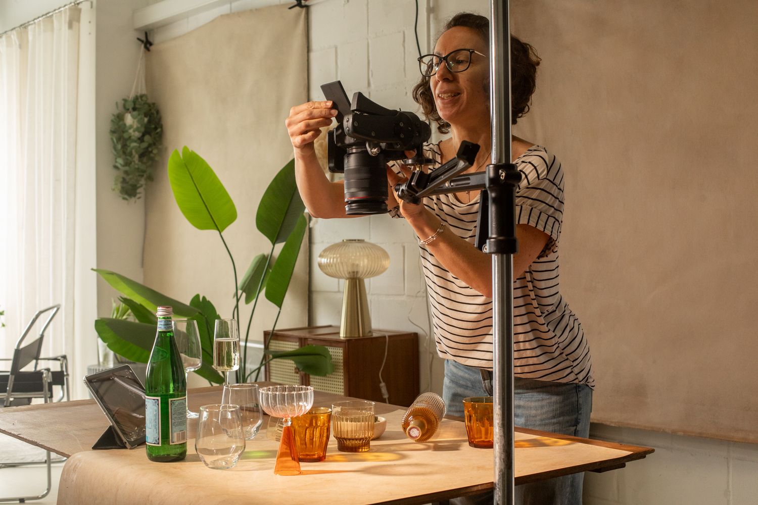 Woman photographer adjusting camera on tripod over table with glassware and bottles in bright studio.