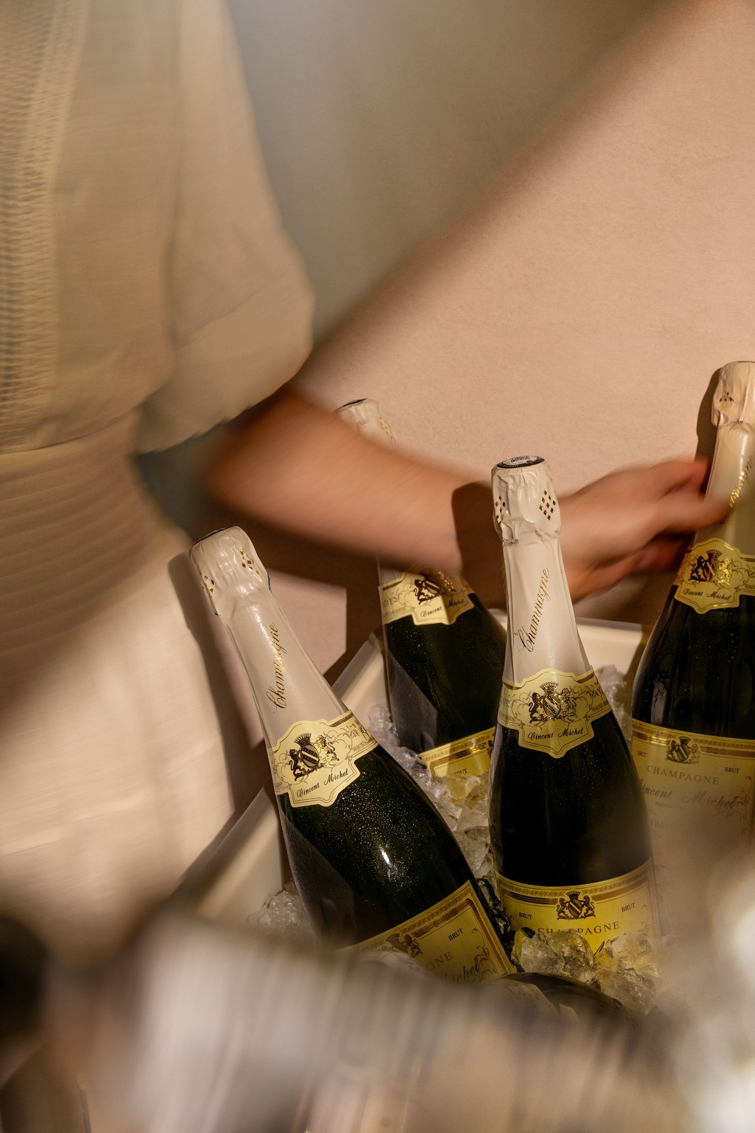 Person in white dress reaching for champagne bottles arranged on a table at an elegant celebration event.