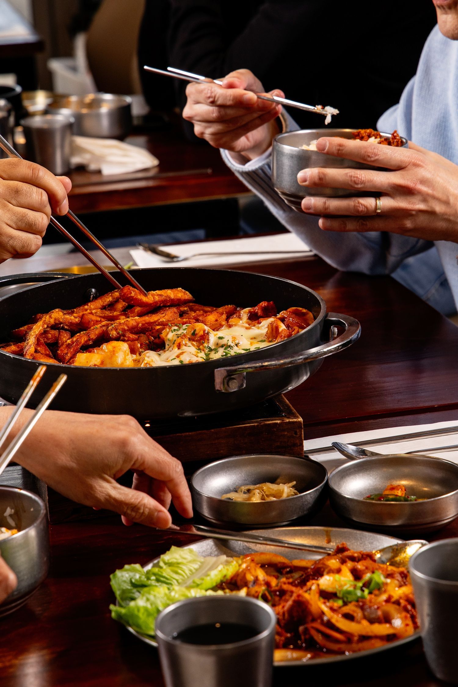 People sharing Korean dishes with chopsticks around a table with spicy stir-fry, side dishes, and dipping sauces.