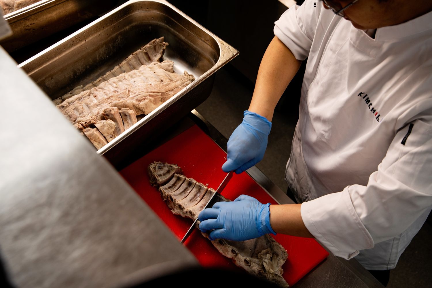 Chef in white uniform and blue gloves filleting fresh fish on a red cutting board in a professional kitchen.