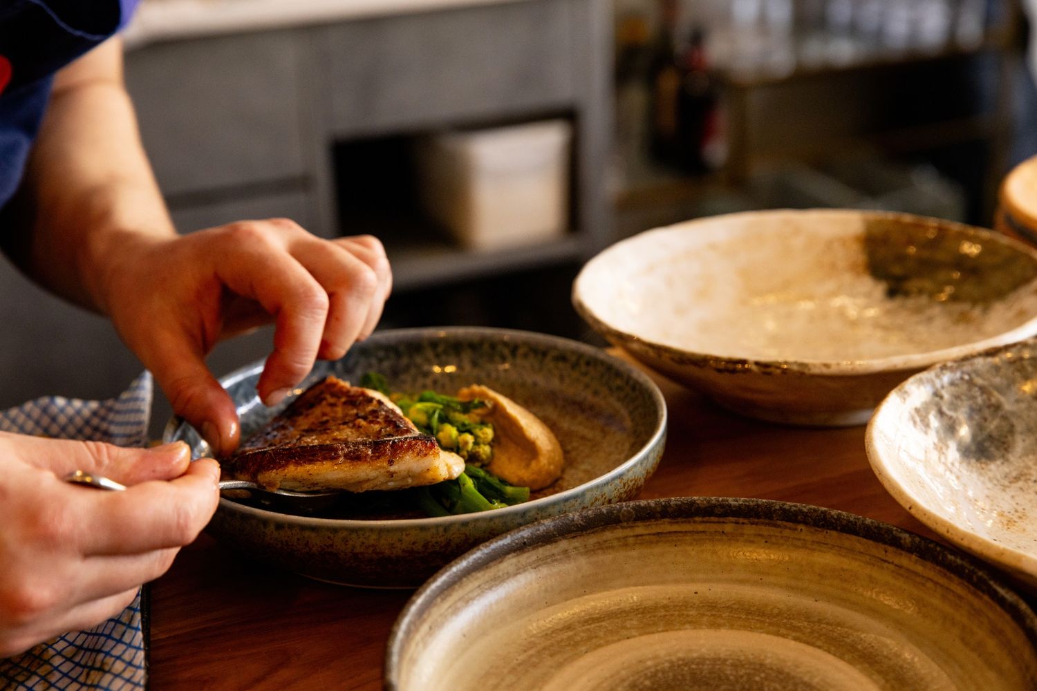 Chef plating a gourmet fish dish with greens on a ceramic plate in a restaurant kitchen.