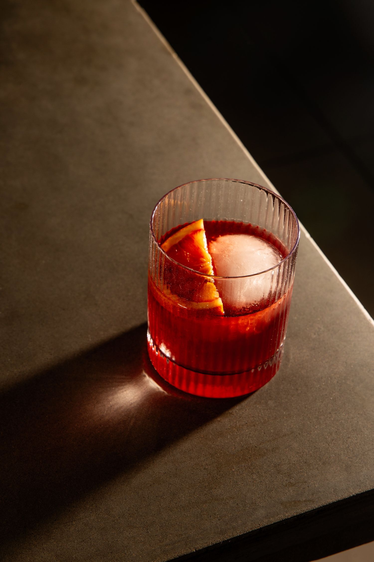 A red cocktail with a large spherical ice cube and orange garnish in a crystal glass casting dramatic shadows.
