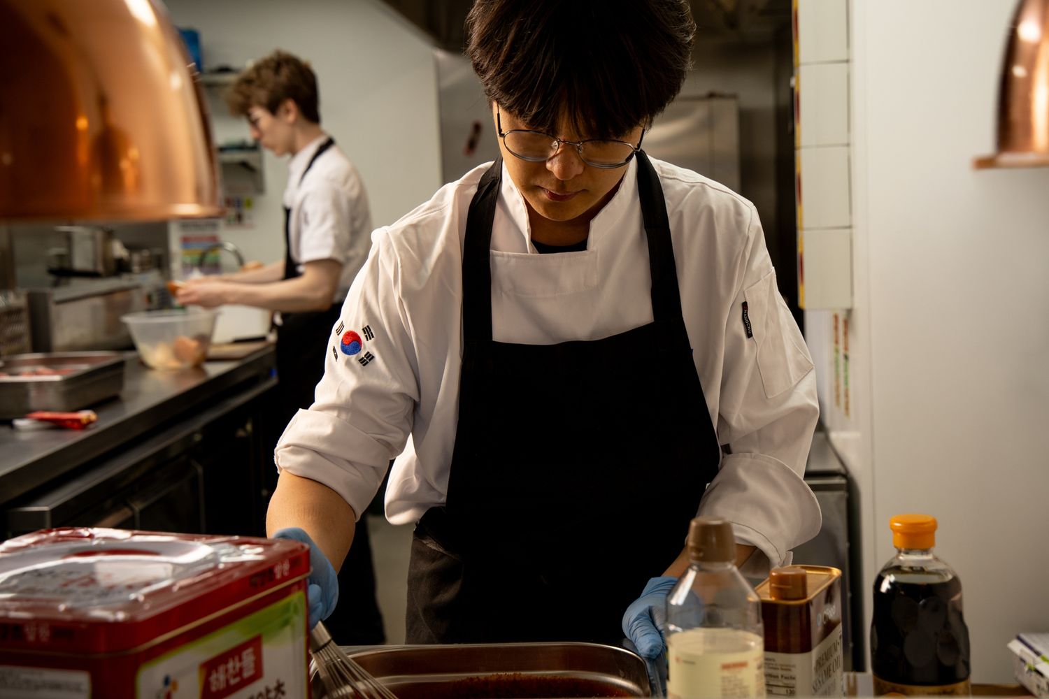 Korean chef in black apron prepares food in a professional restaurant kitchen with another cook working in background.