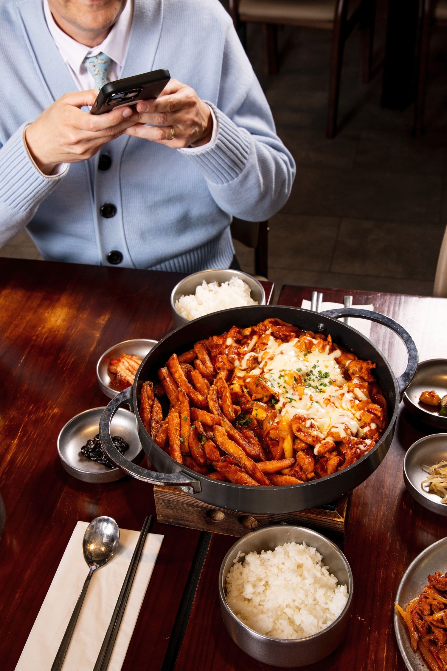 Person photographing a Korean tteokbokki hot pot with melted cheese, rice bowls, and banchan side dishes on a wooden table.