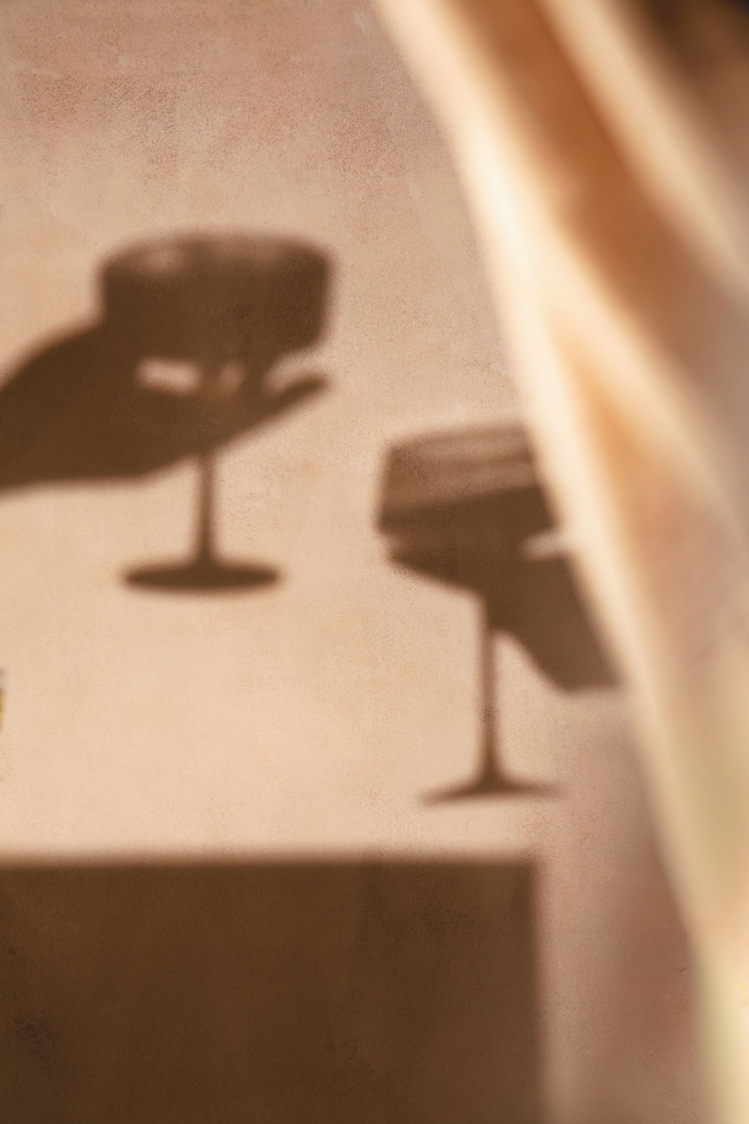 Soft shadows of two wine glasses cast on a warm beige wall with draped fabric in golden light.
