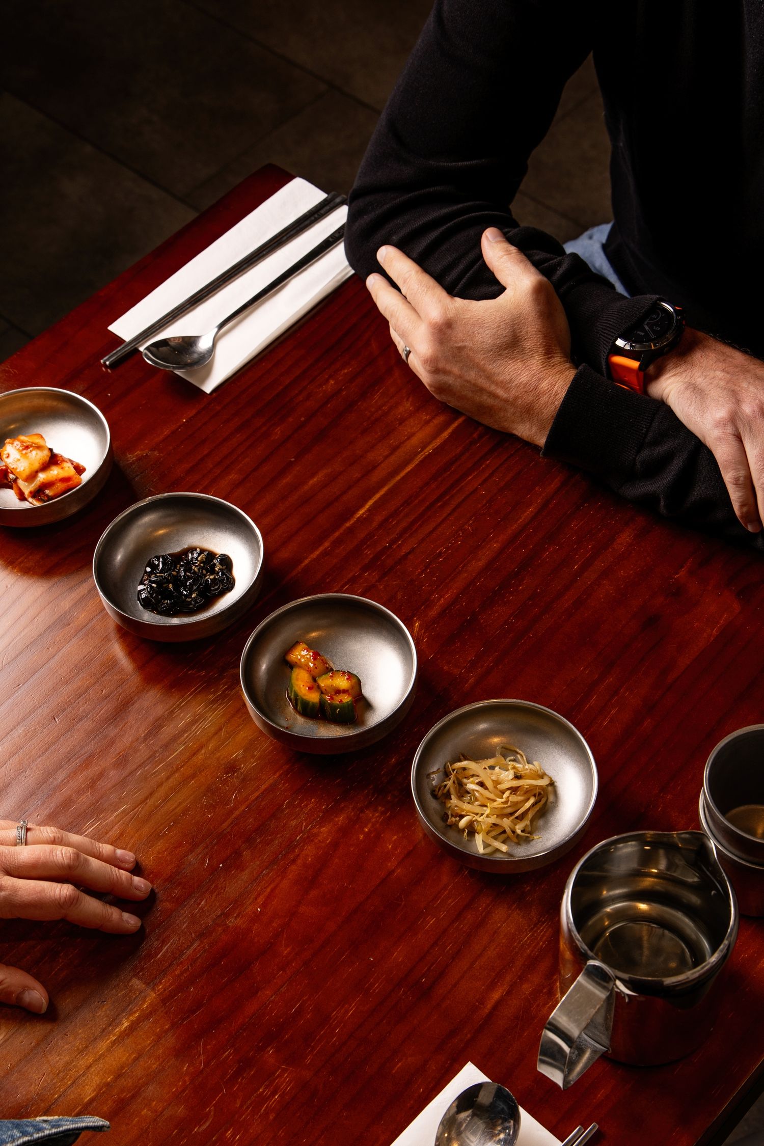 Overhead view of Korean banchan side dishes in silver bowls on a wooden table with two people dining.