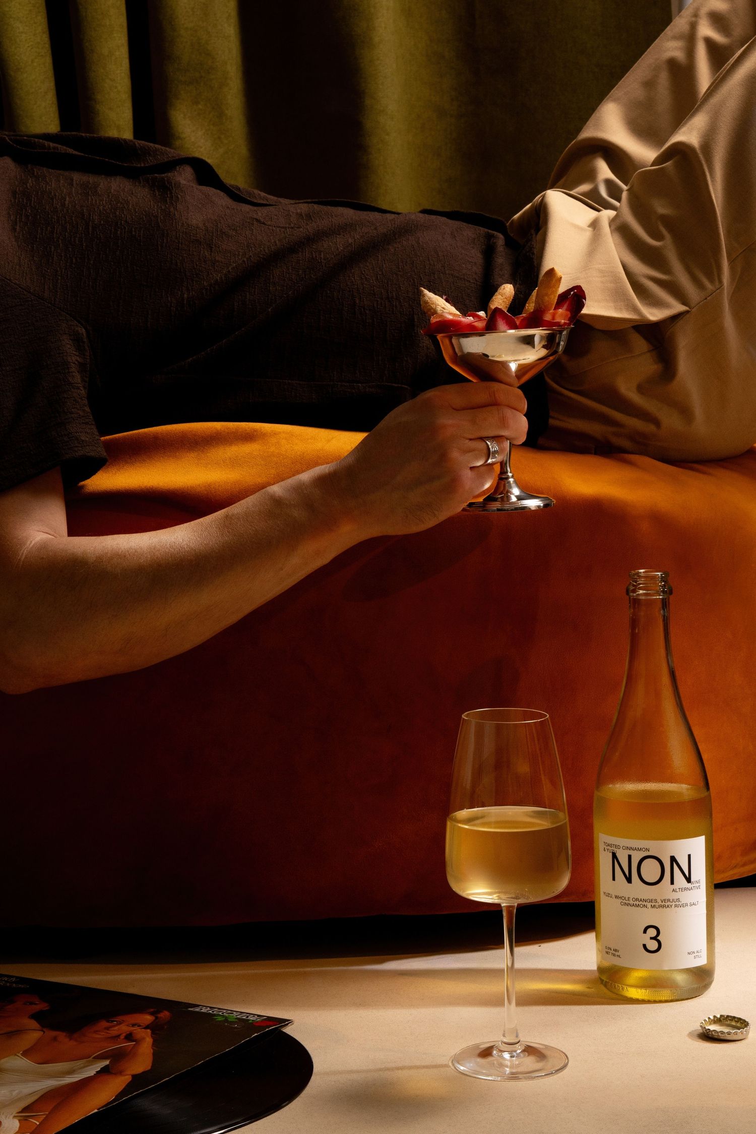 Person holding a garnished cocktail glass beside a bottle of NON 3 non-alcoholic beverage and a wine glass on a table.