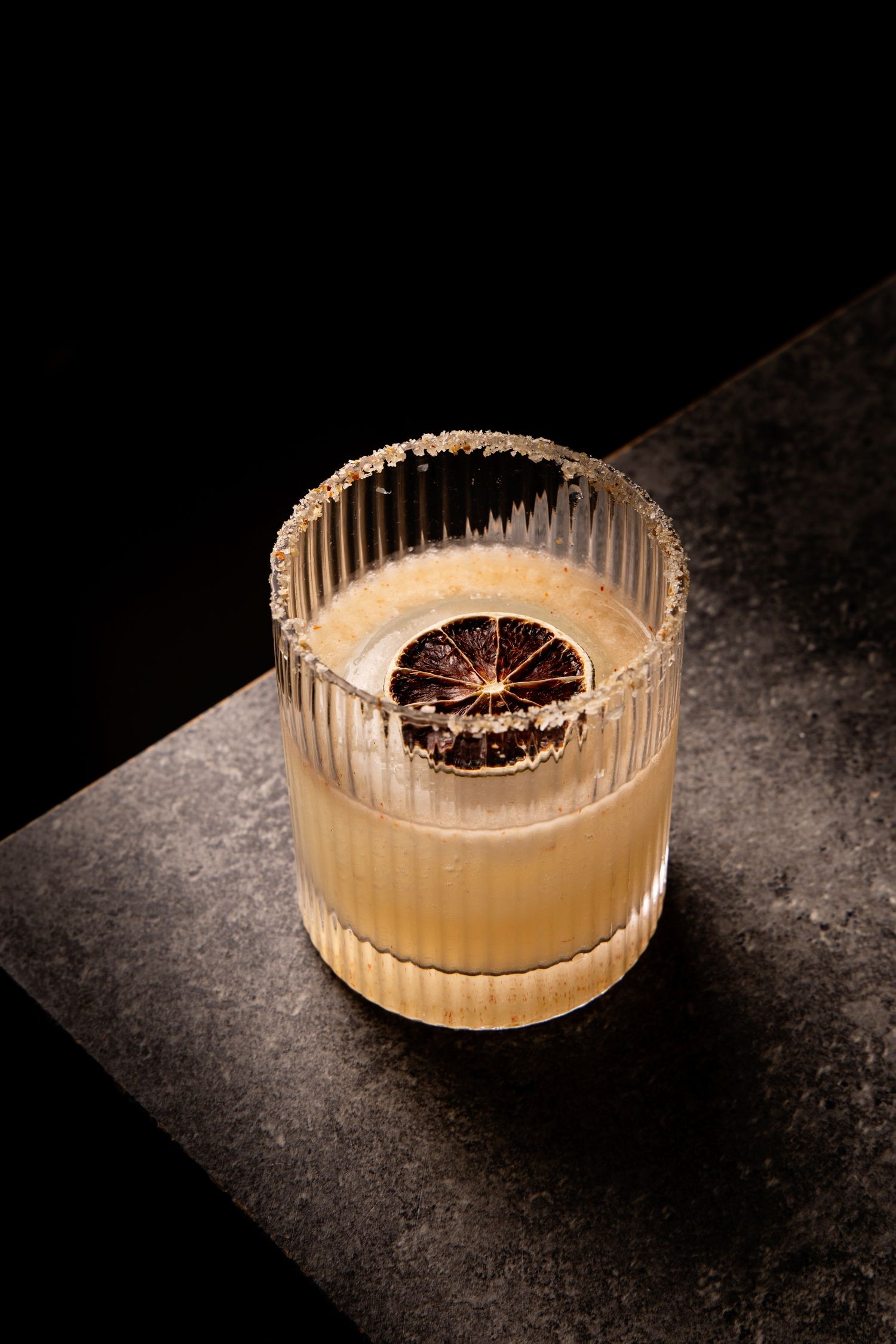 A craft cocktail with citrus garnish served in a ribbed crystal glass on a dark surface with dramatic lighting.