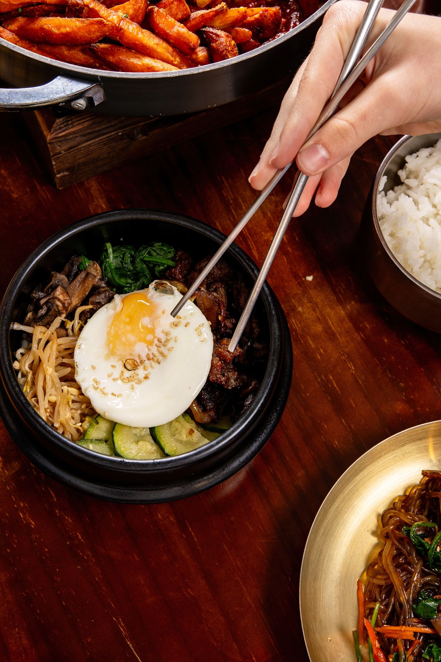 Hand using chopsticks to pick up a fried egg in a Korean bibimbap bowl, with rice and tteokbokki nearby.