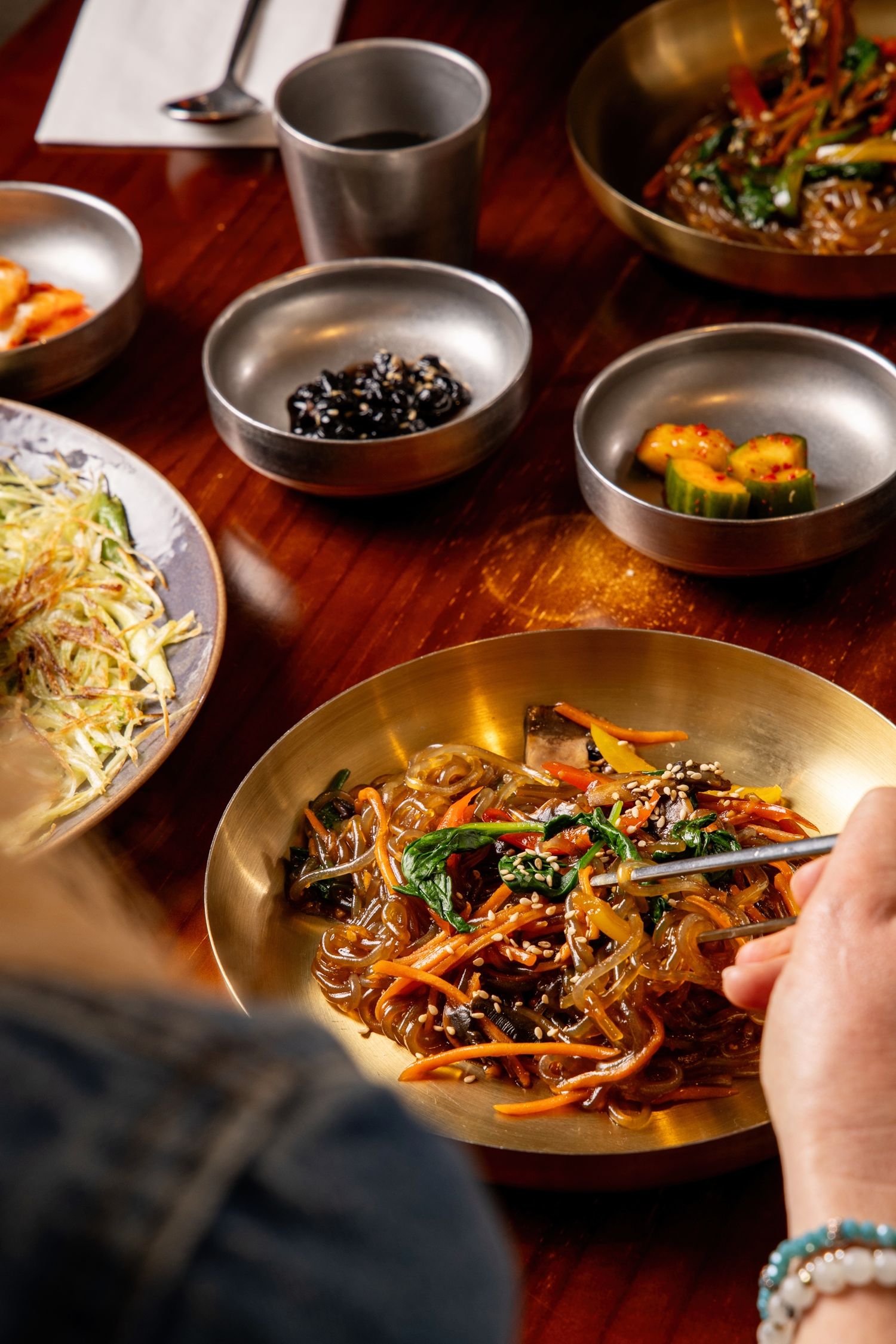 Korean japchae glass noodles with vegetables served in gold bowl alongside traditional banchan side dishes.