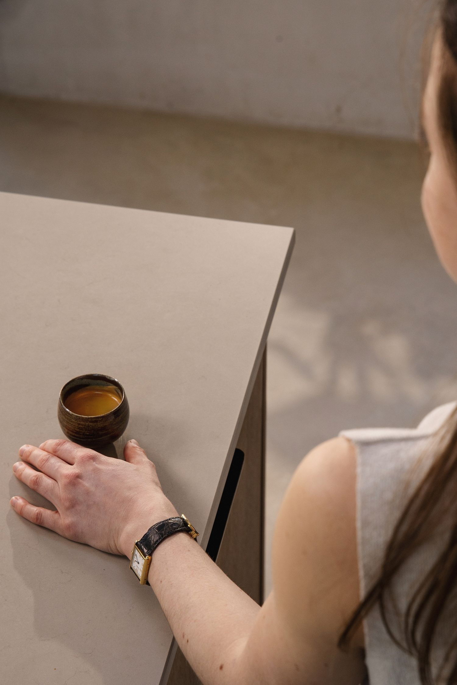 Person sitting at desk holding small glass cup of espresso with watch on wrist and pen on table.