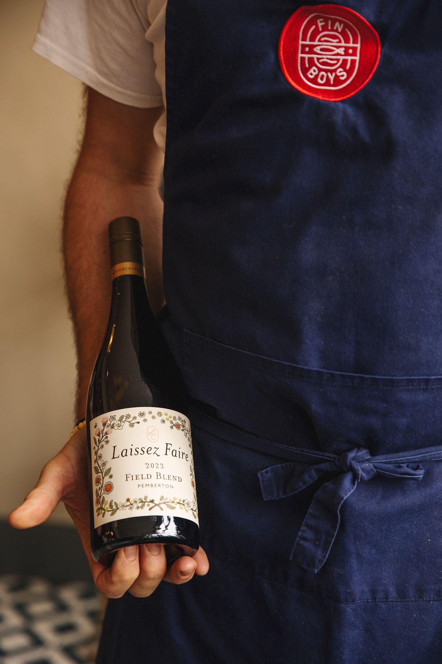 Person in blue apron holds wine bottles showing labels against neutral background.