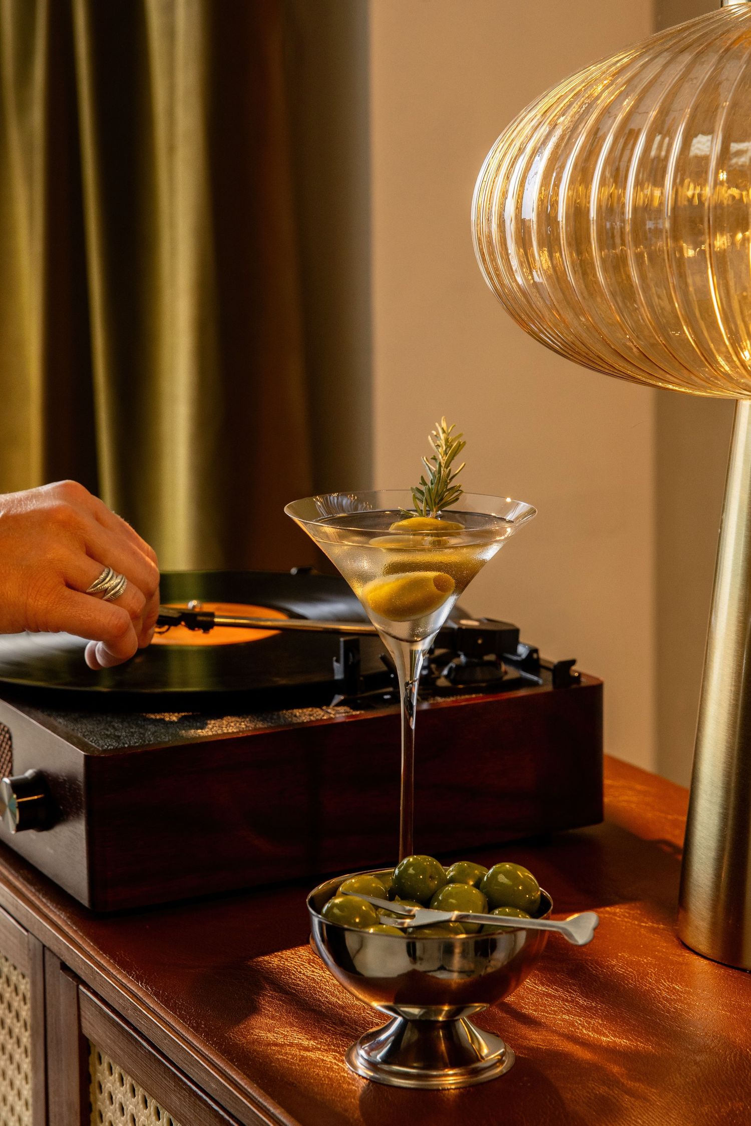 Elegant dirty martini with olives beside a vinyl record player and golden lamp on a wooden sideboard.