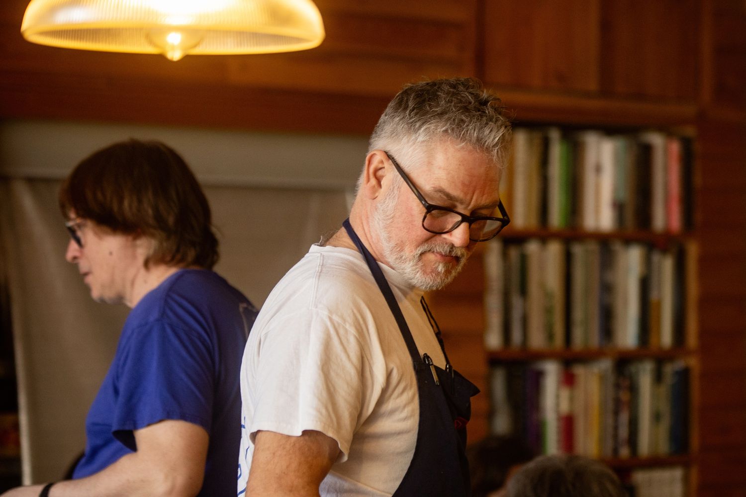 Two chefs in aprons work in a cozy kitchen with bookshelves, focused on food preparation under warm lighting.