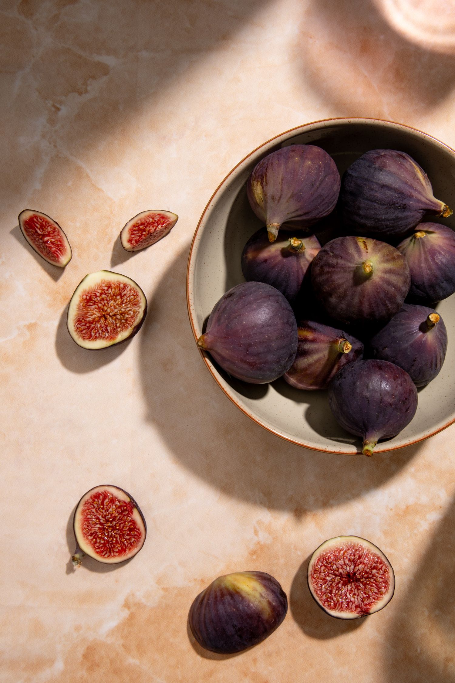 Fresh purple figs in a ceramic bowl with sliced figs on a marble surface in warm sunlight.