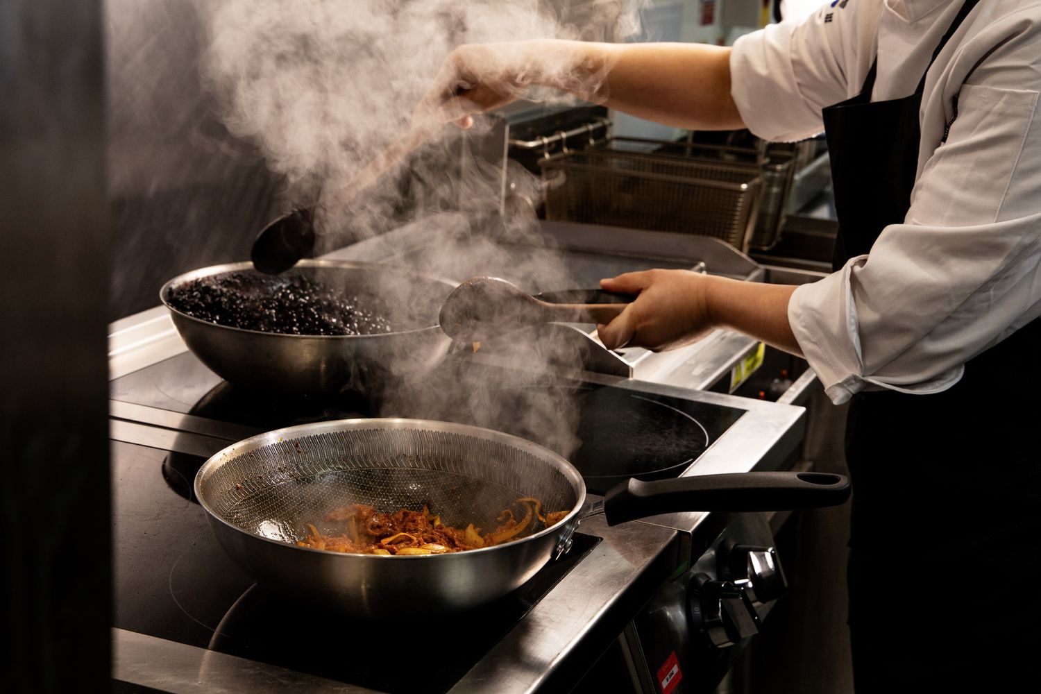 Chef cooking in a professional kitchen with steaming woks and pans on an induction stove.