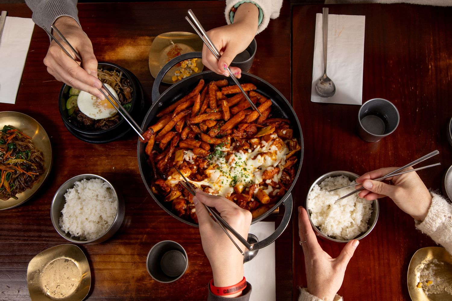 Overhead view of people sharing Korean tteokbokki in a black pan with rice bowls and side dishes on a wooden table.