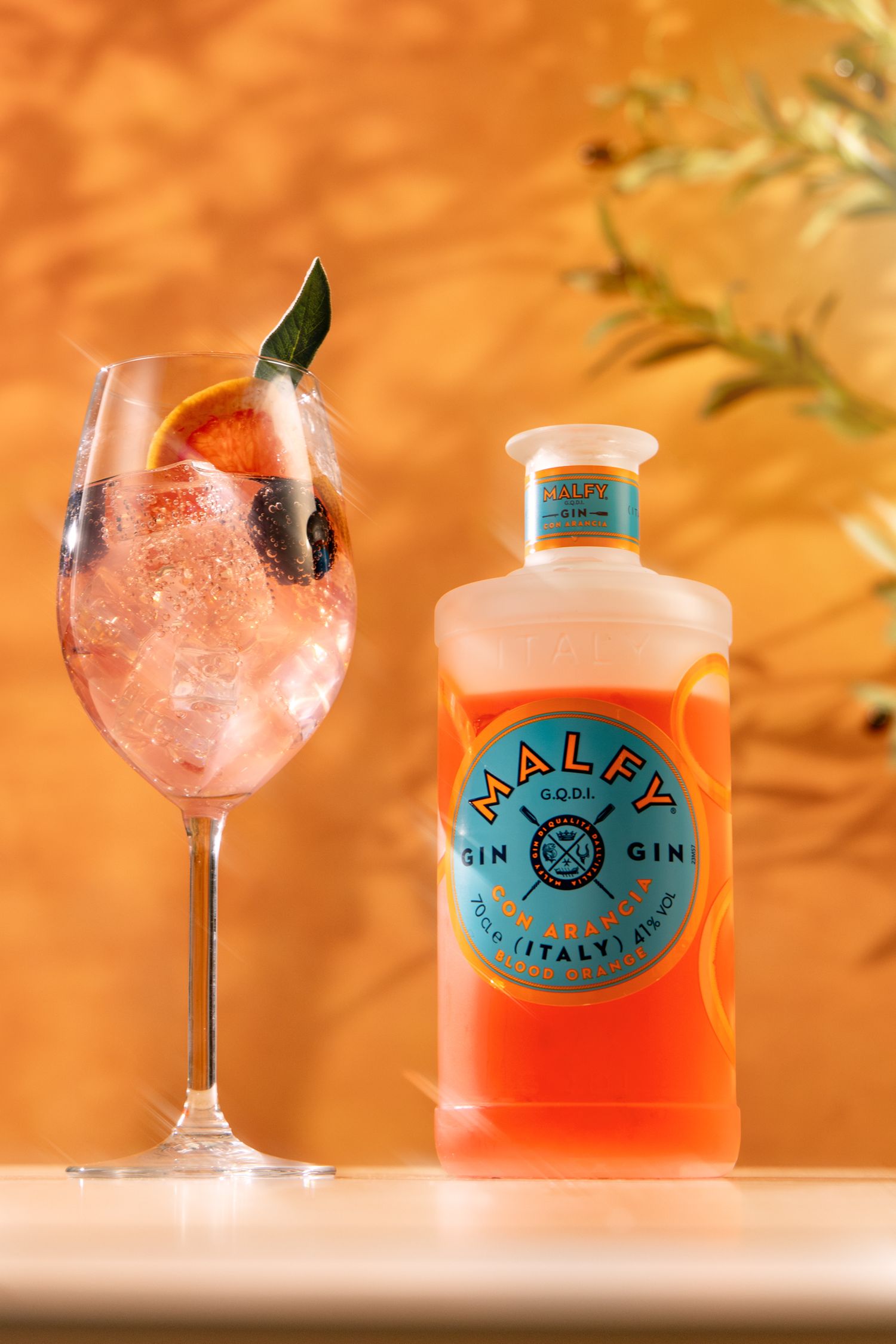 A pink cocktail with garnish in a wine glass next to a bottle of Malfy gin against an orange-lit background.