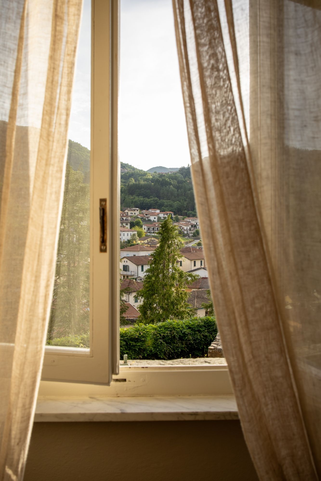 Sunlit room interior with flowing sheer curtains and mountain village view through windows.