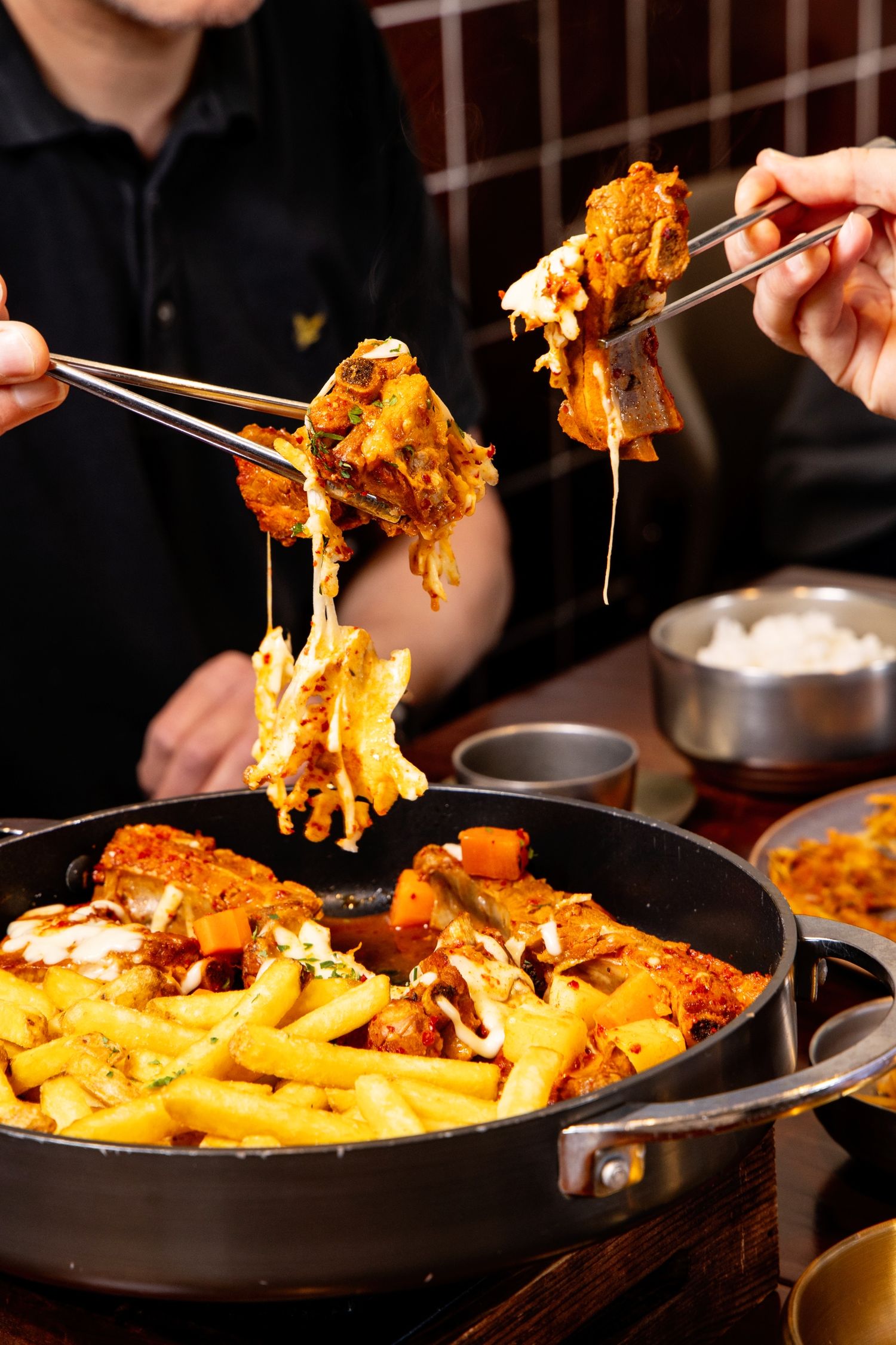 Two people sharing a Korean cheese buldak dish with fries, pulling melted cheese strings with tongs from a sizzling pan.