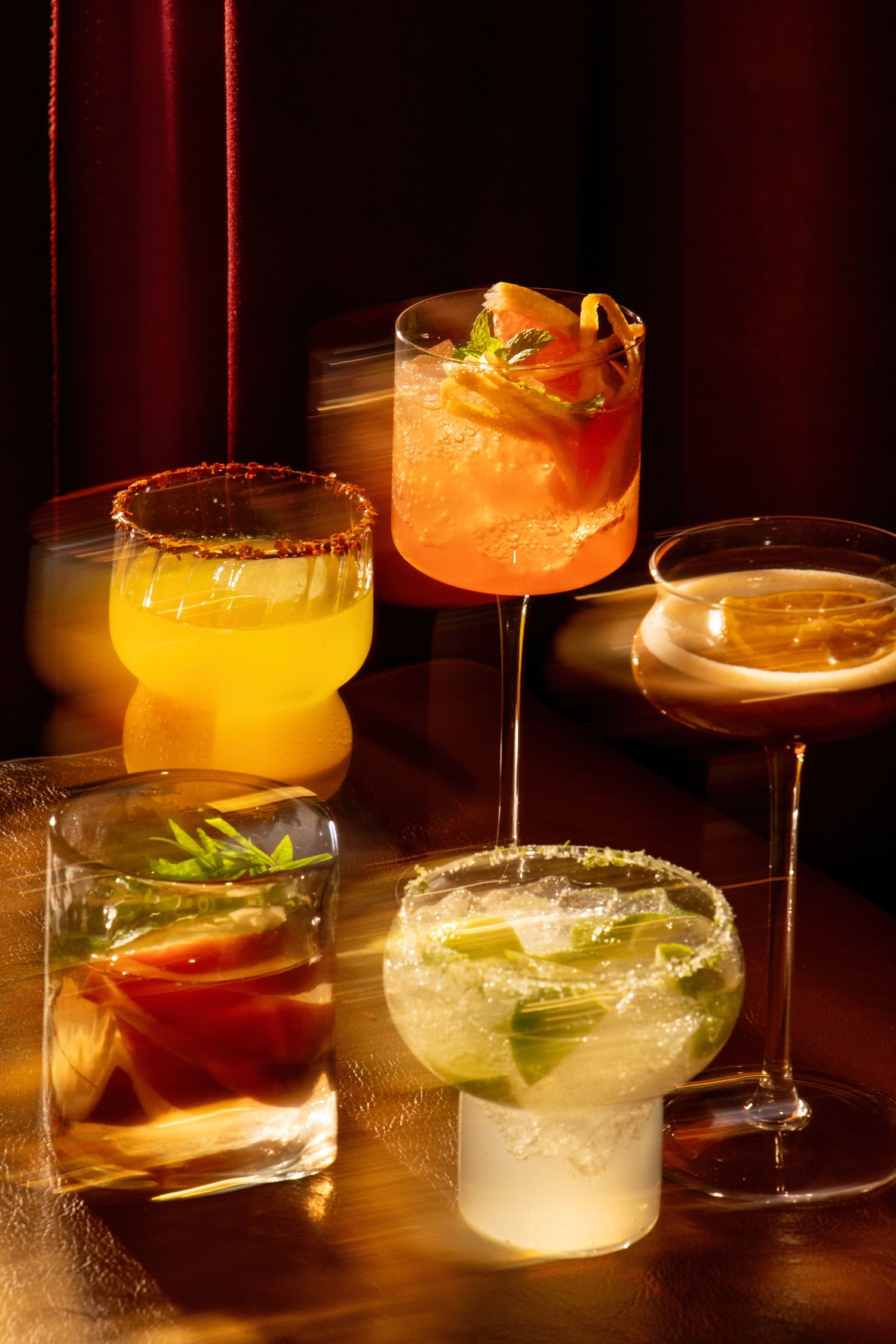 Five colorful cocktails in various glasses on a wooden table, illuminated by warm golden light in a moody bar setting.