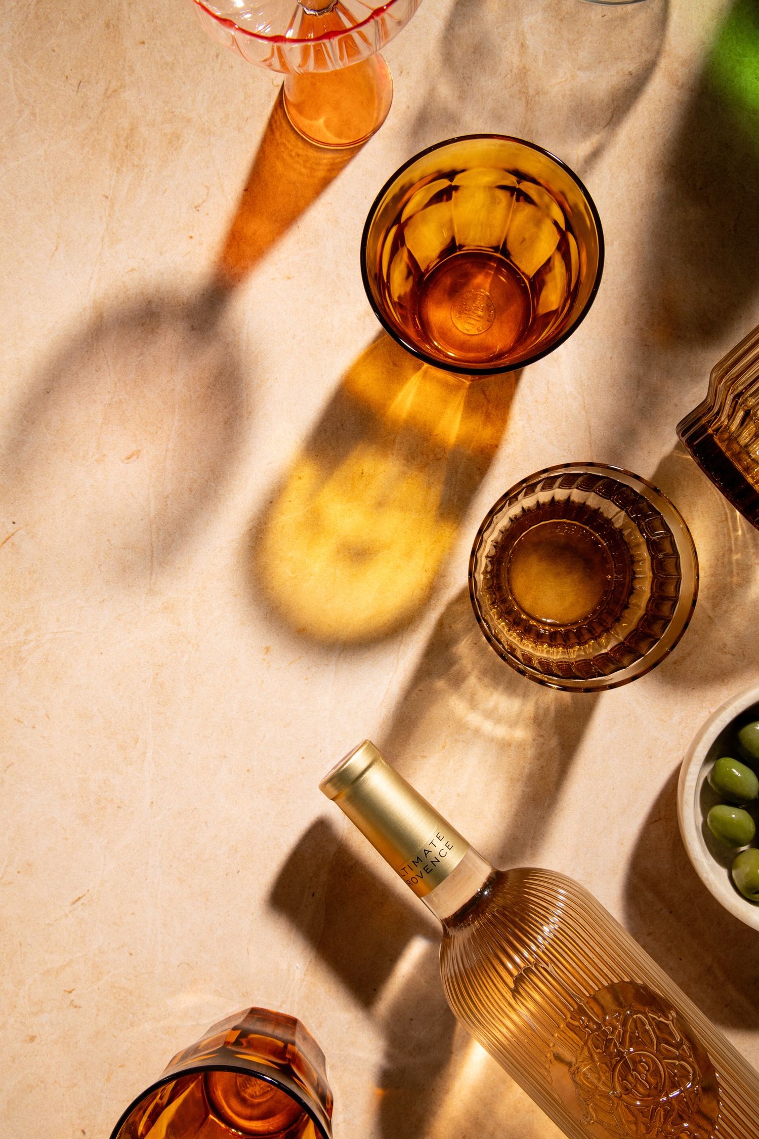 Amber glassware and wine bottle casting warm golden shadows on a beige surface with olives nearby.