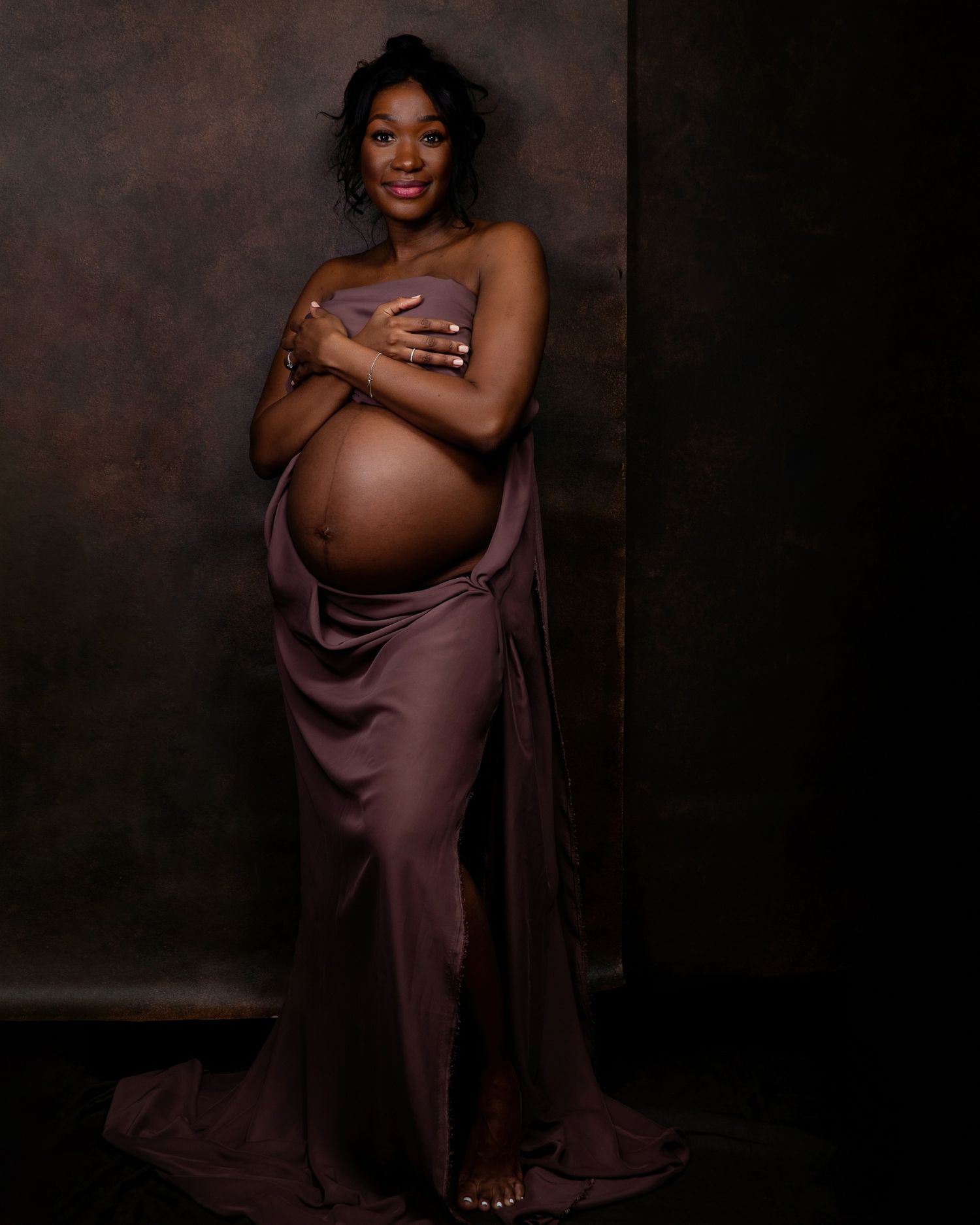 Maternity photoshoot featuring a woman in a flowing mauve dress against a dark backdrop.