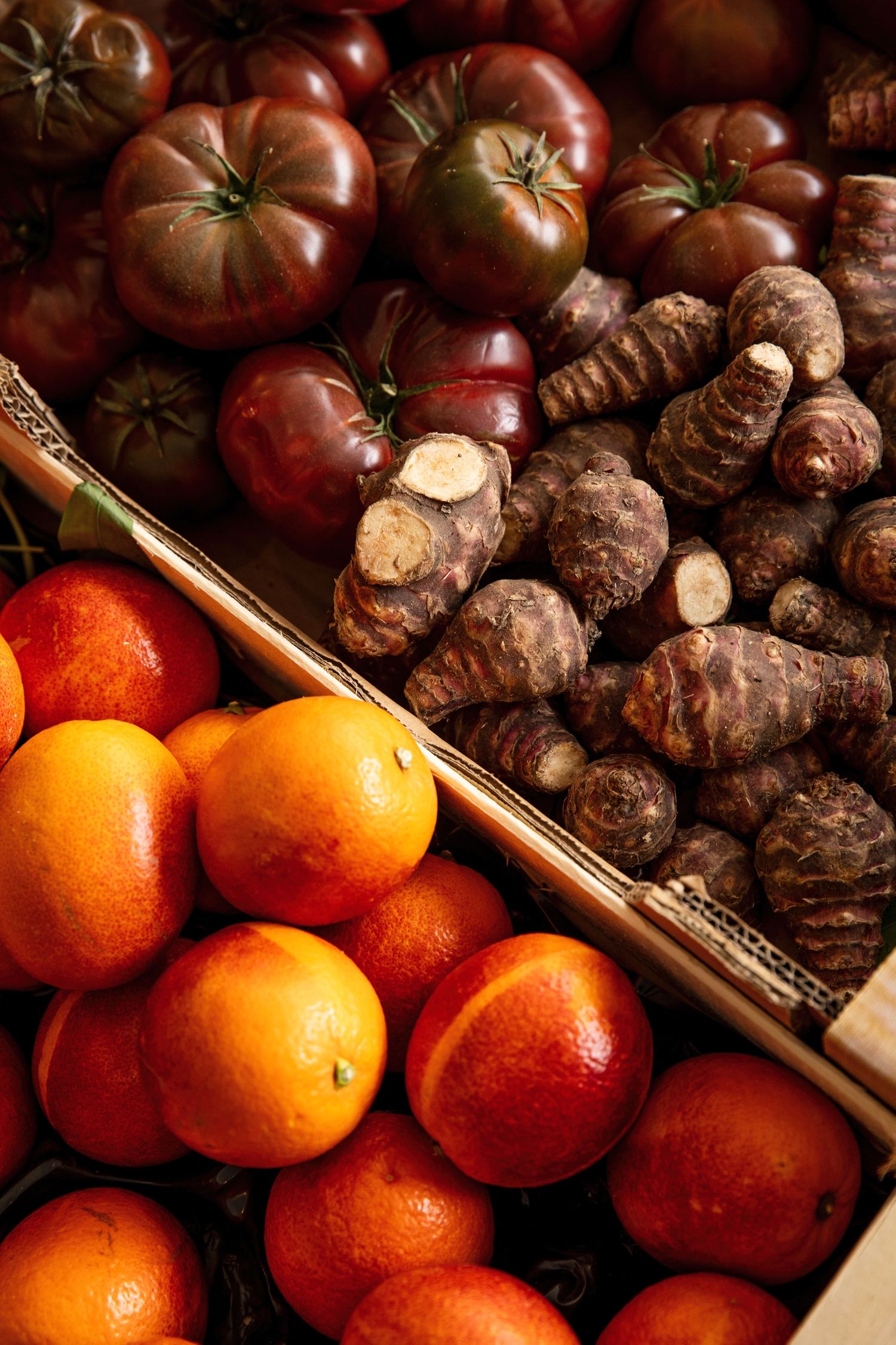 Fresh oranges and dark red tomatoes arranged with brown Jerusalem artichokes on a wooden surface in warm lighting.