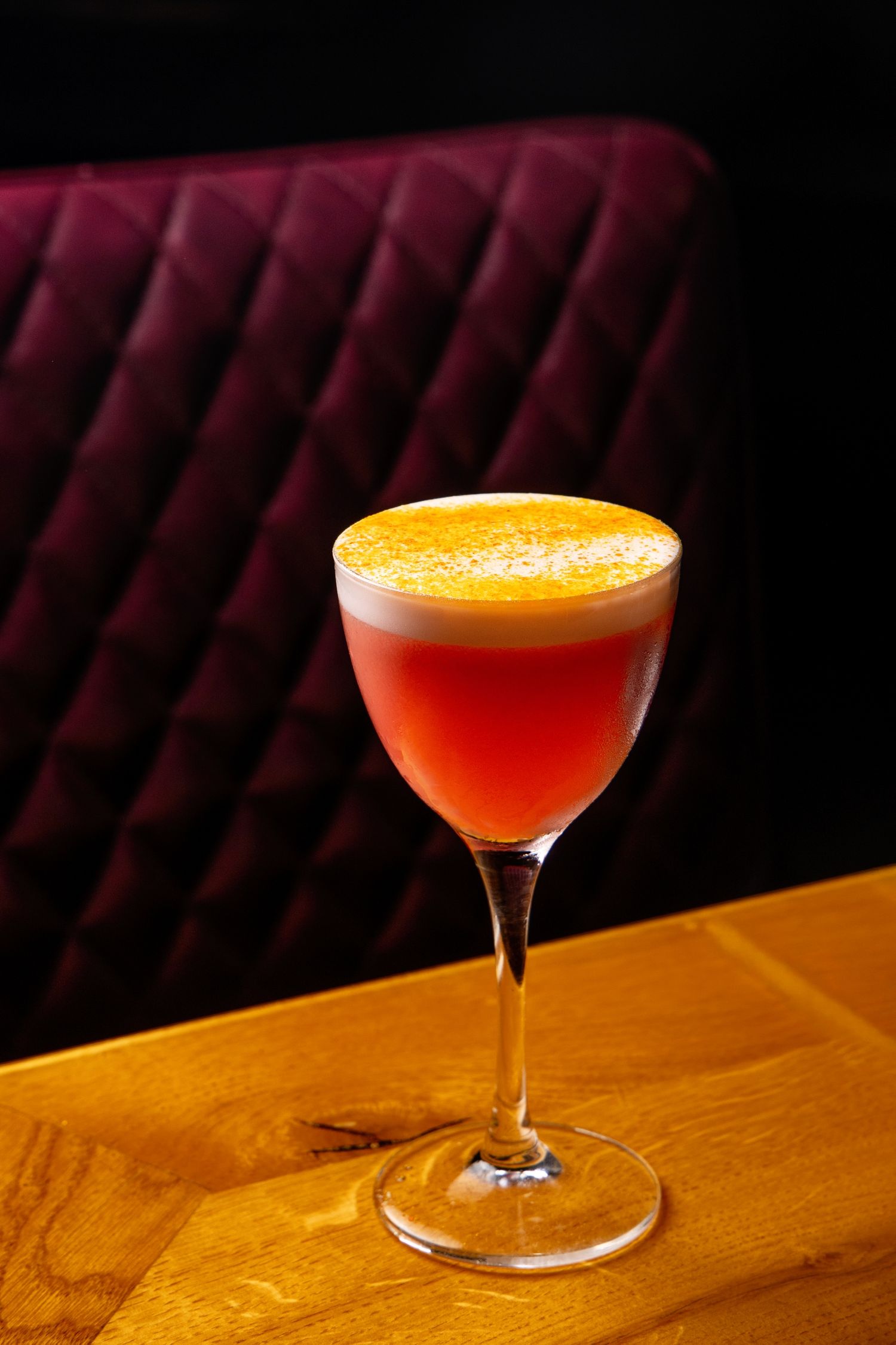A sequence of cocktail glasses filled with orange and red drinks on a wooden bar surface in dim lighting.