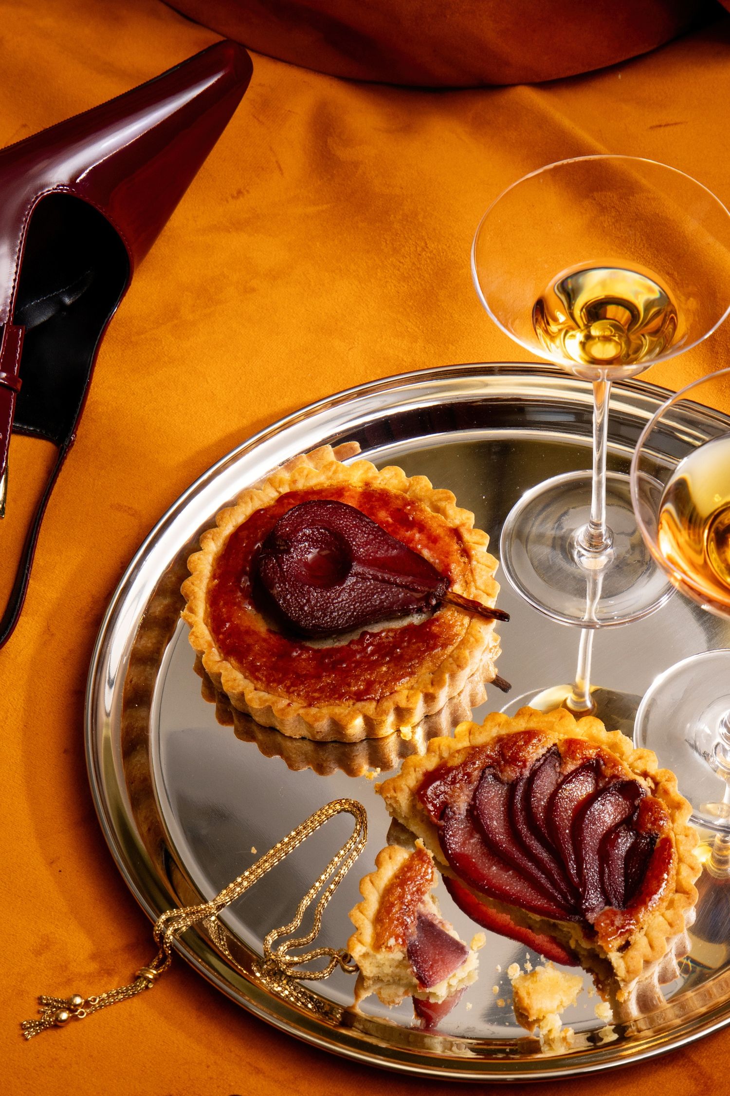 Elegant pear tarts on silver tray with white wine glass and burgundy heels on golden fabric backdrop.
