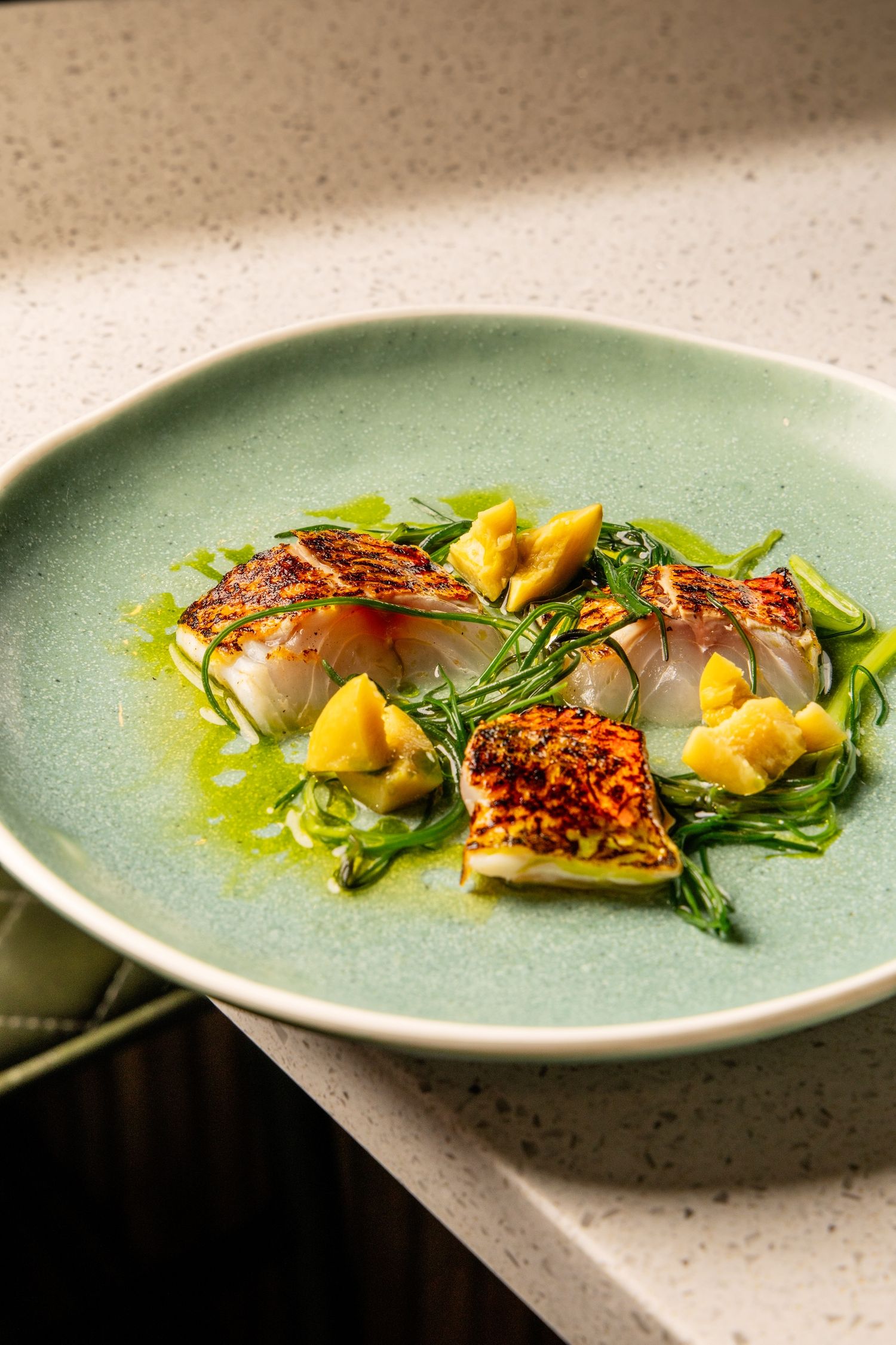 Pan-seared fish fillets served on a bed of greens with mango chunks and herb oil on a turquoise ceramic plate.