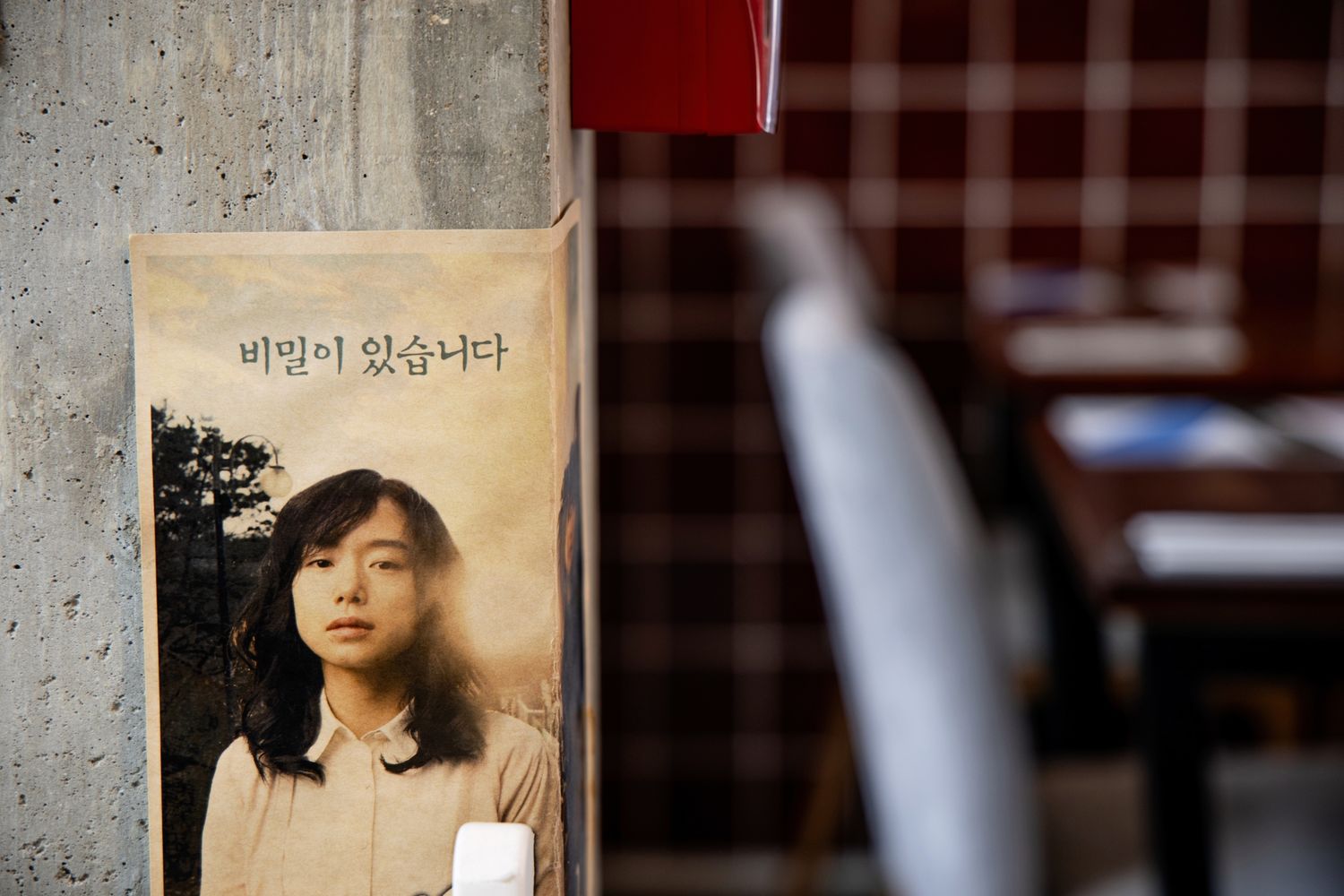 Korean poster with a young woman's portrait and Hangul text displayed on a concrete wall near a checkered curtain.