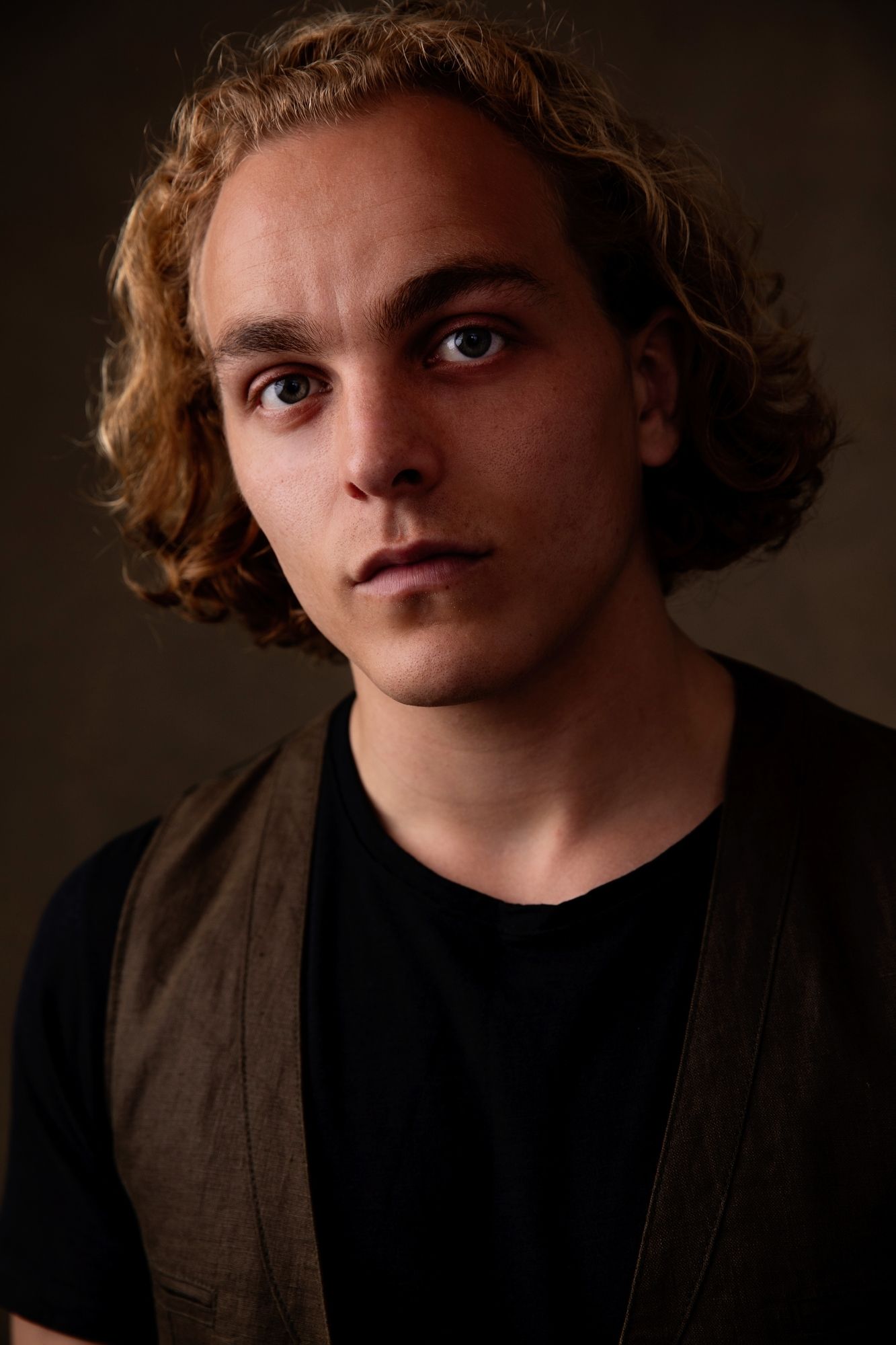Moody portrait of a young person with wavy hair wearing dark clothing against shadowy background.
