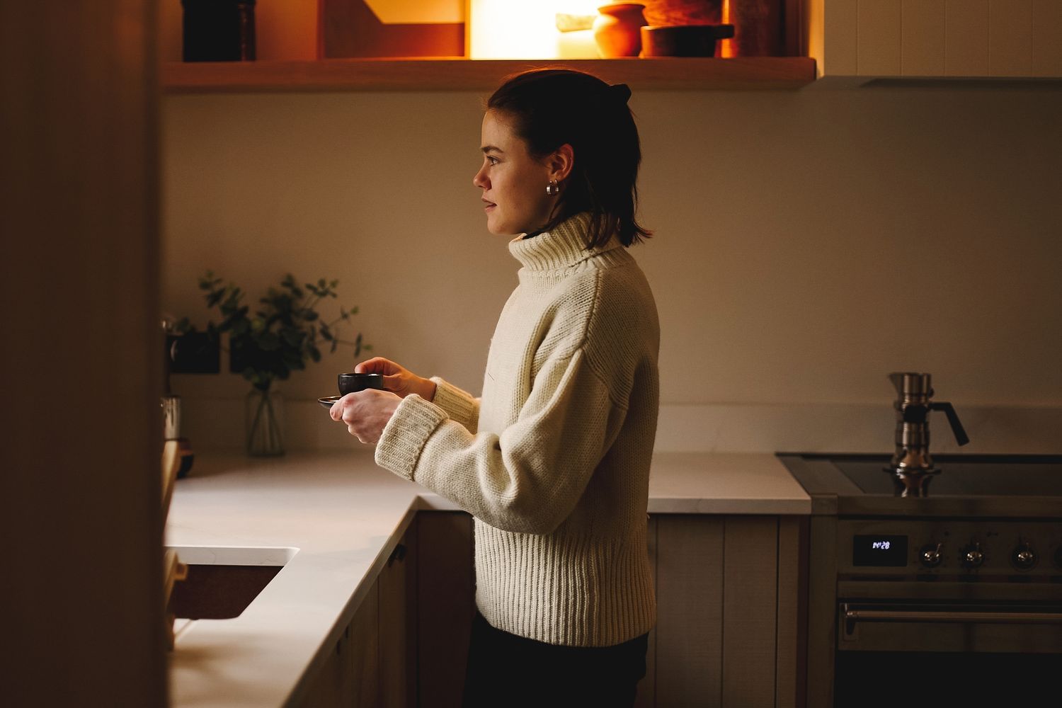 Person in cozy sweater relaxing at home with a coffee in hands with warm lighting in the background.