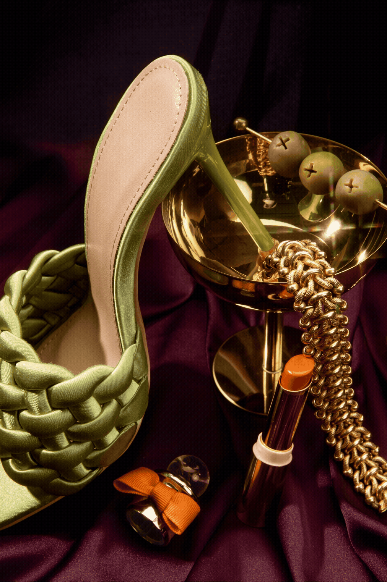 Green braided high heel mule shoe beside gold martini glass, lipstick, chain bracelet on purple silk fabric.