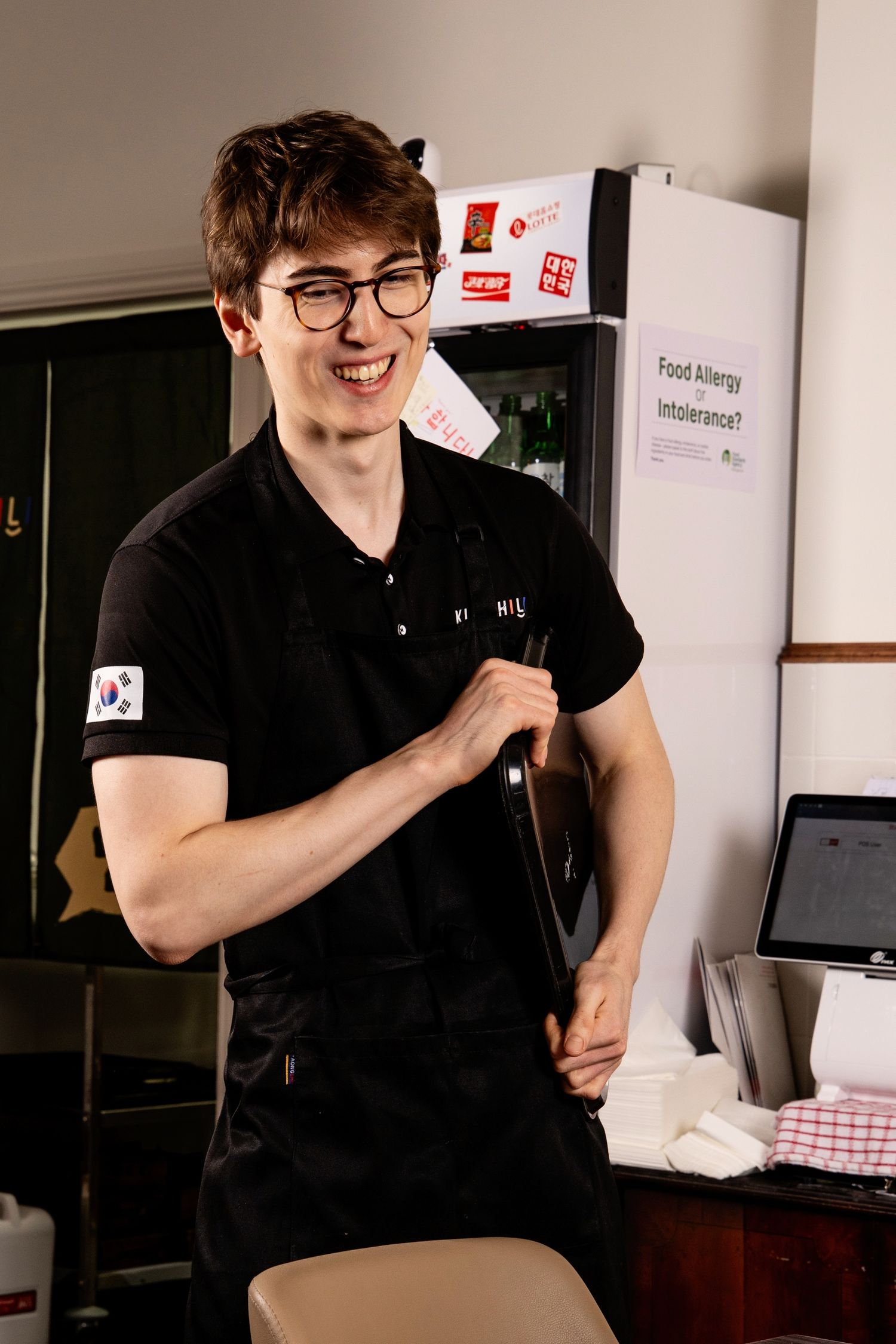 Young smiling waiter in black polo with South Korean flag patch holding a wine bottle in a restaurant.