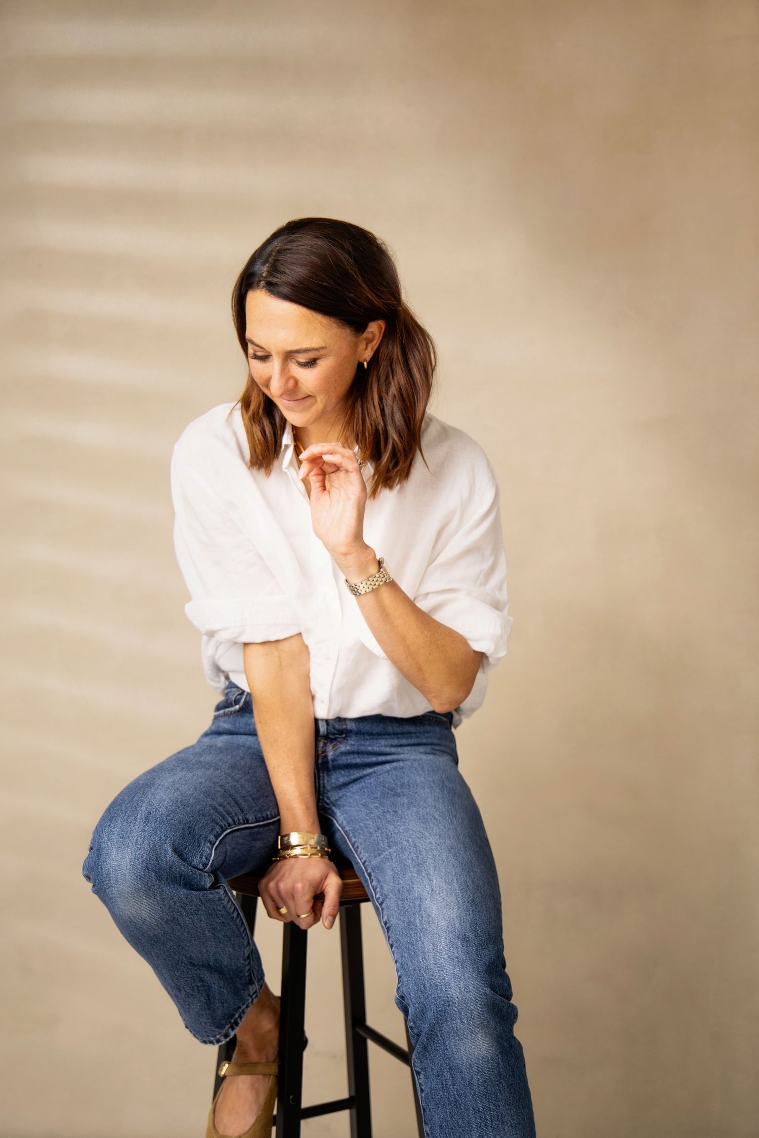 Person in white t-shirt and blue jeans sits casually on dark stool against neutral background.