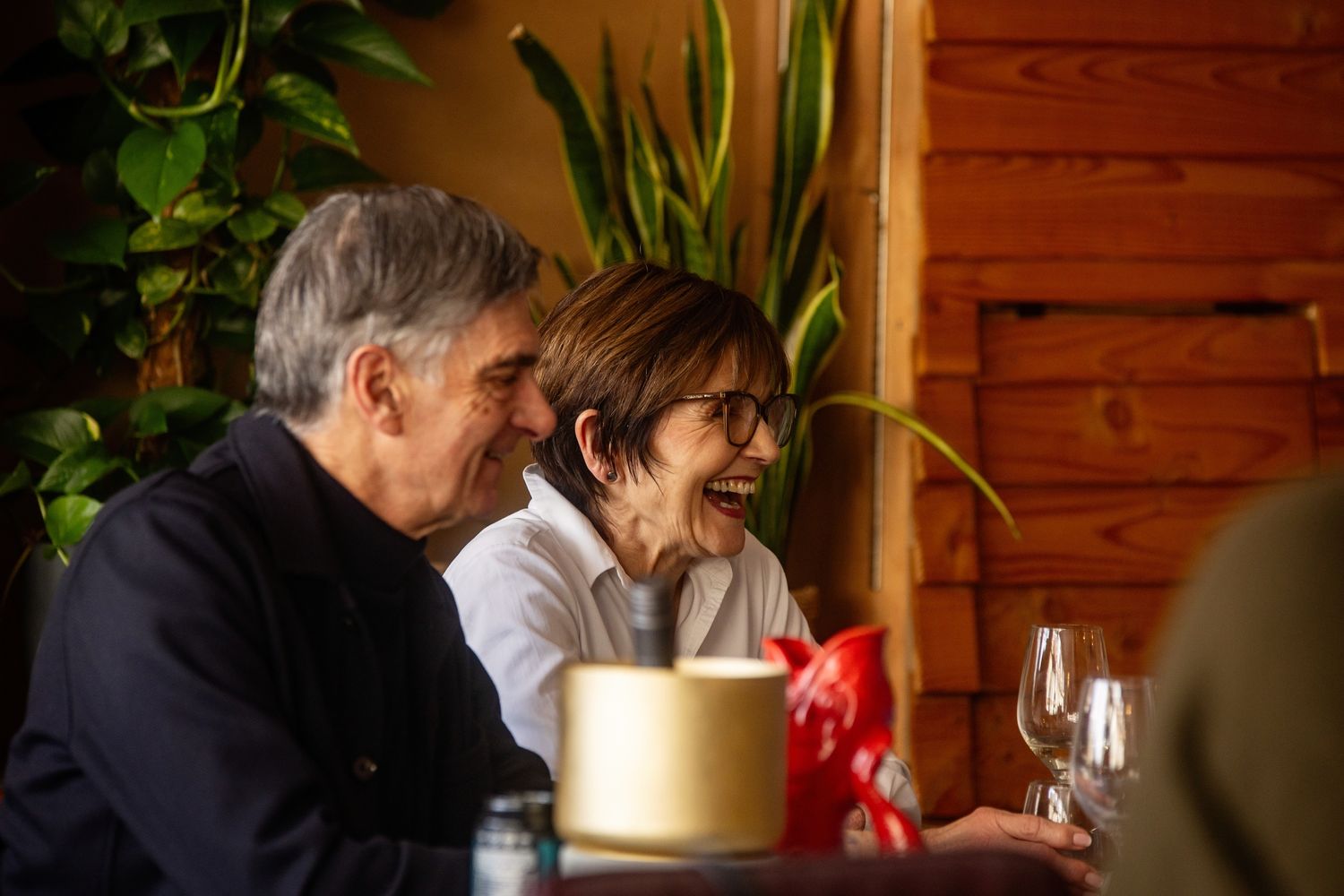 An elderly couple enjoying a cozy conversation over a drink in a rustic setting with potted plants.