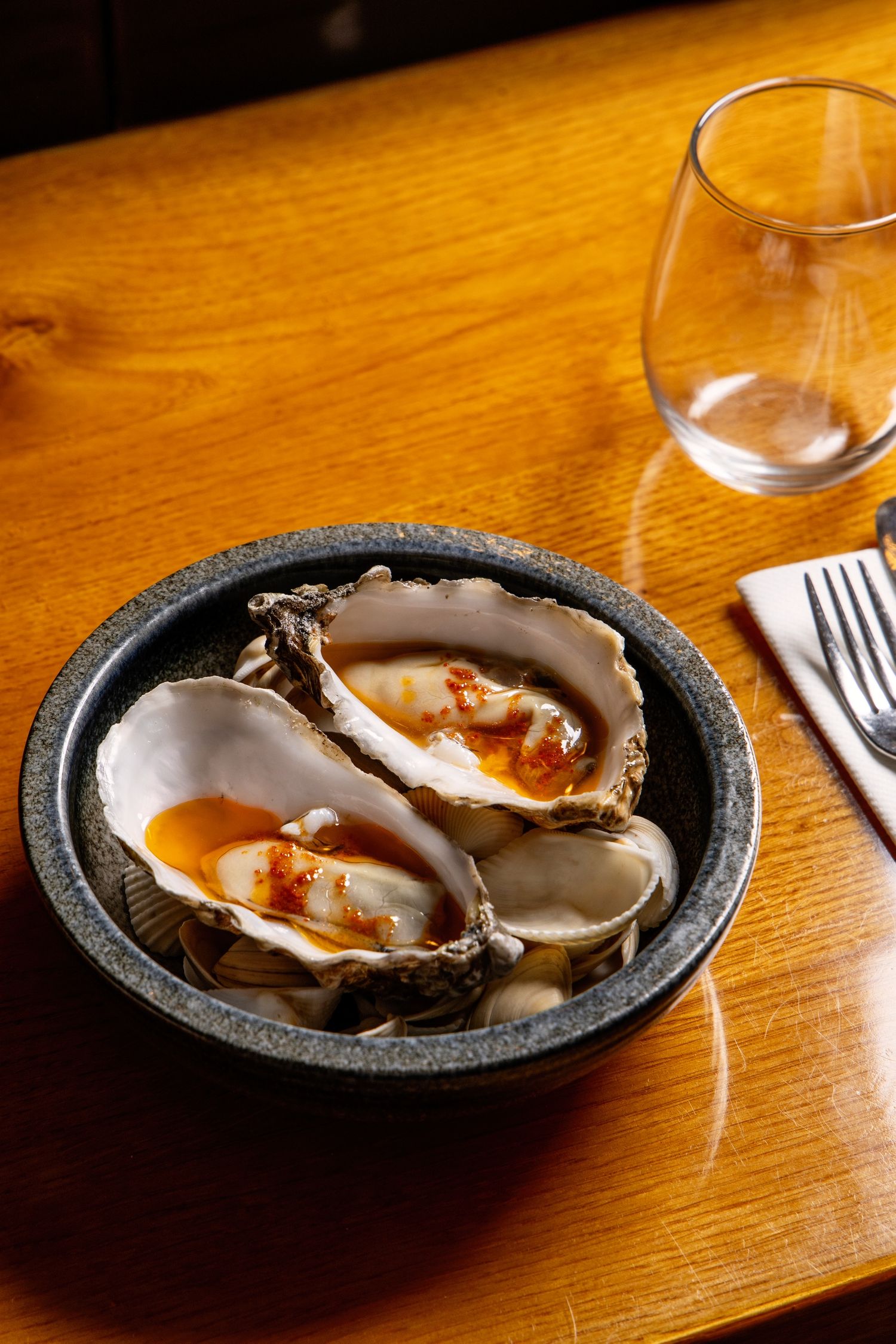Fresh oysters on dark stone plates sit on a wooden table with wine glasses in the background.