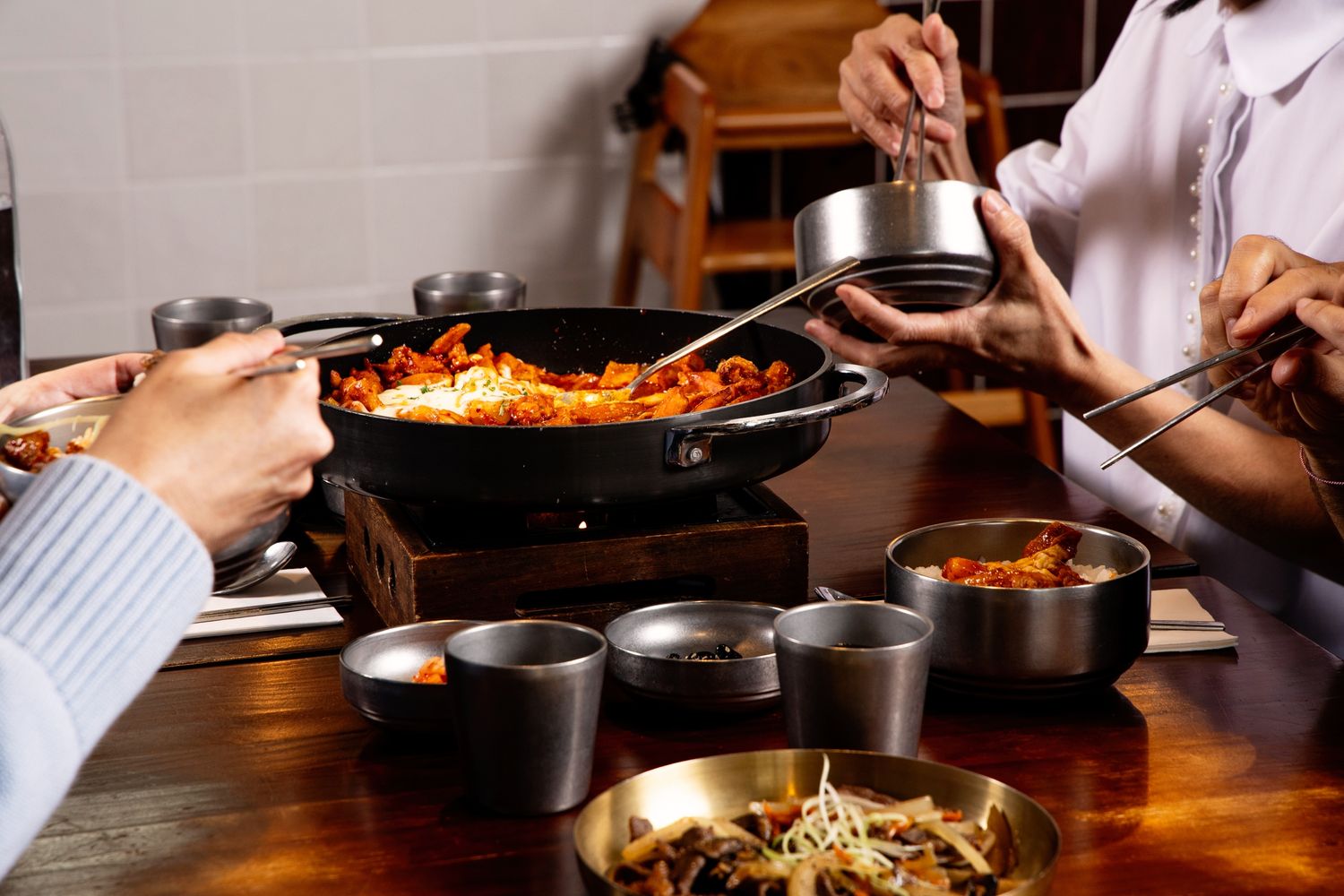 People sharing a spicy Korean stir-fry dish in a black pan with metal bowls and chopsticks on a wooden table.