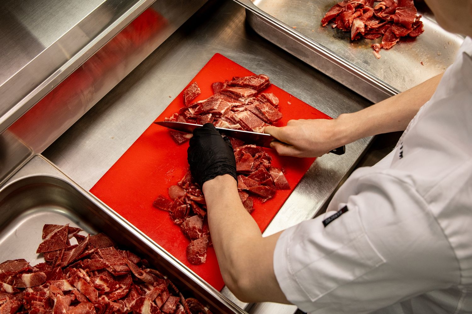 Chef in black gloves chopping fresh red meat on a cutting board in a professional kitchen.