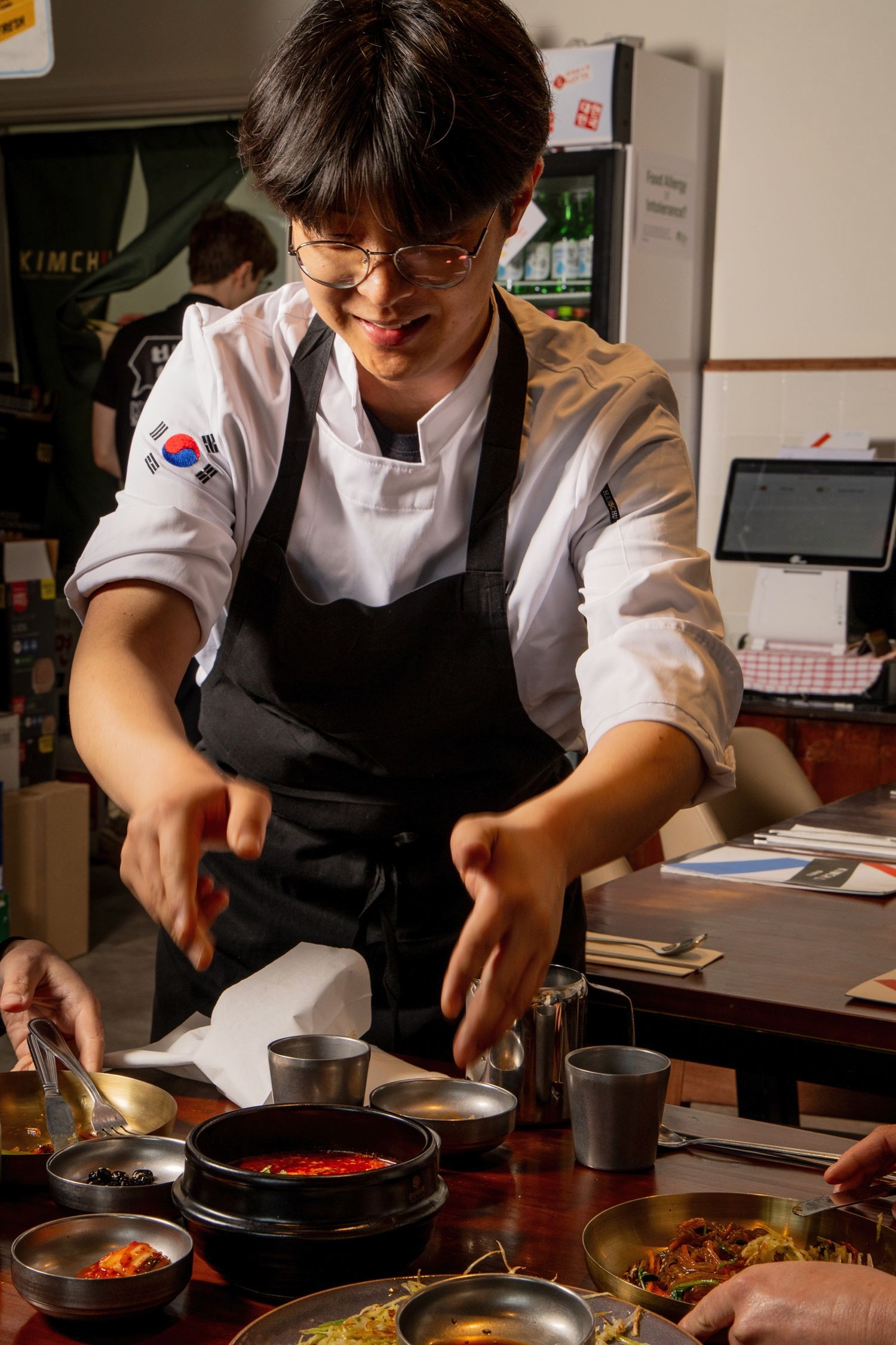Korean chef in white coat and black apron carefully plates a dish with chopsticks in a restaurant kitchen.