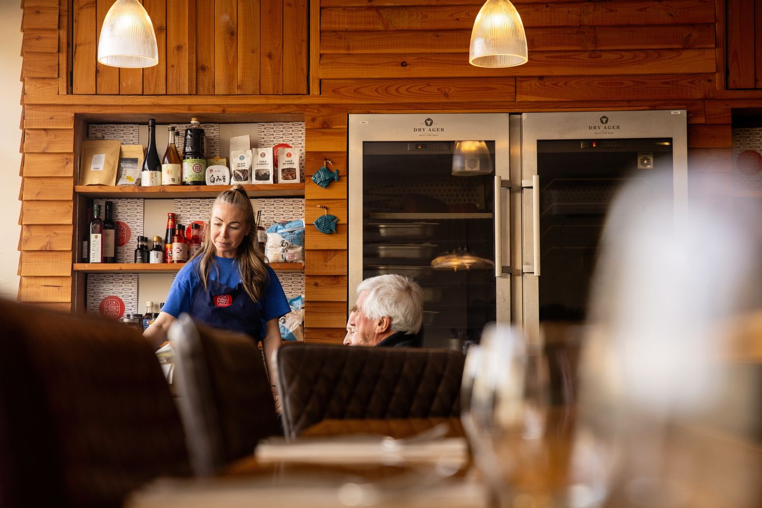 A person sitting in a cozy wooden cabin interior with shelves and appliances visible.