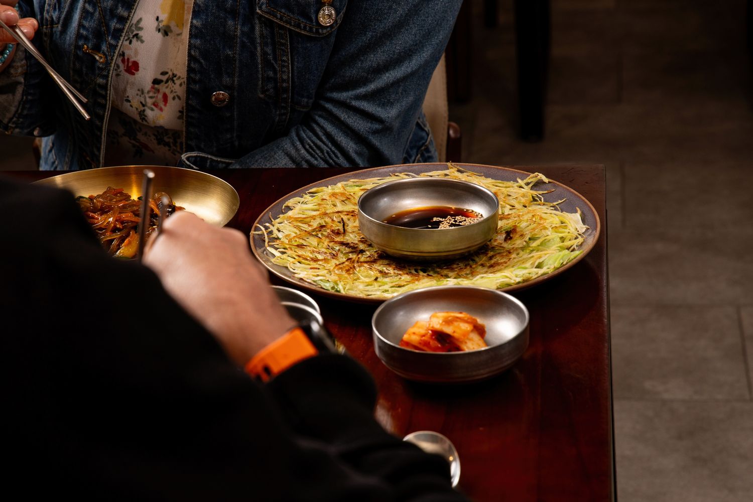 Korean dining table with noodle dish, kimchi, and dipping sauces in silver bowls served on a dark wooden table.