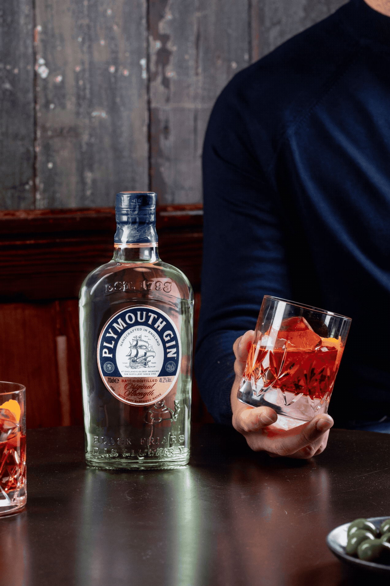 A Plymouth Gin bottle and cocktail glass sit on a dark wooden table with dramatic lighting.
