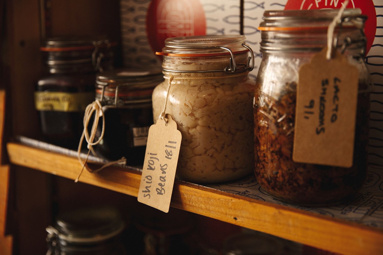 Glass mason jars with handwritten labels containing spices and ingredients sit on a rustic wooden shelf in dim lighting.
