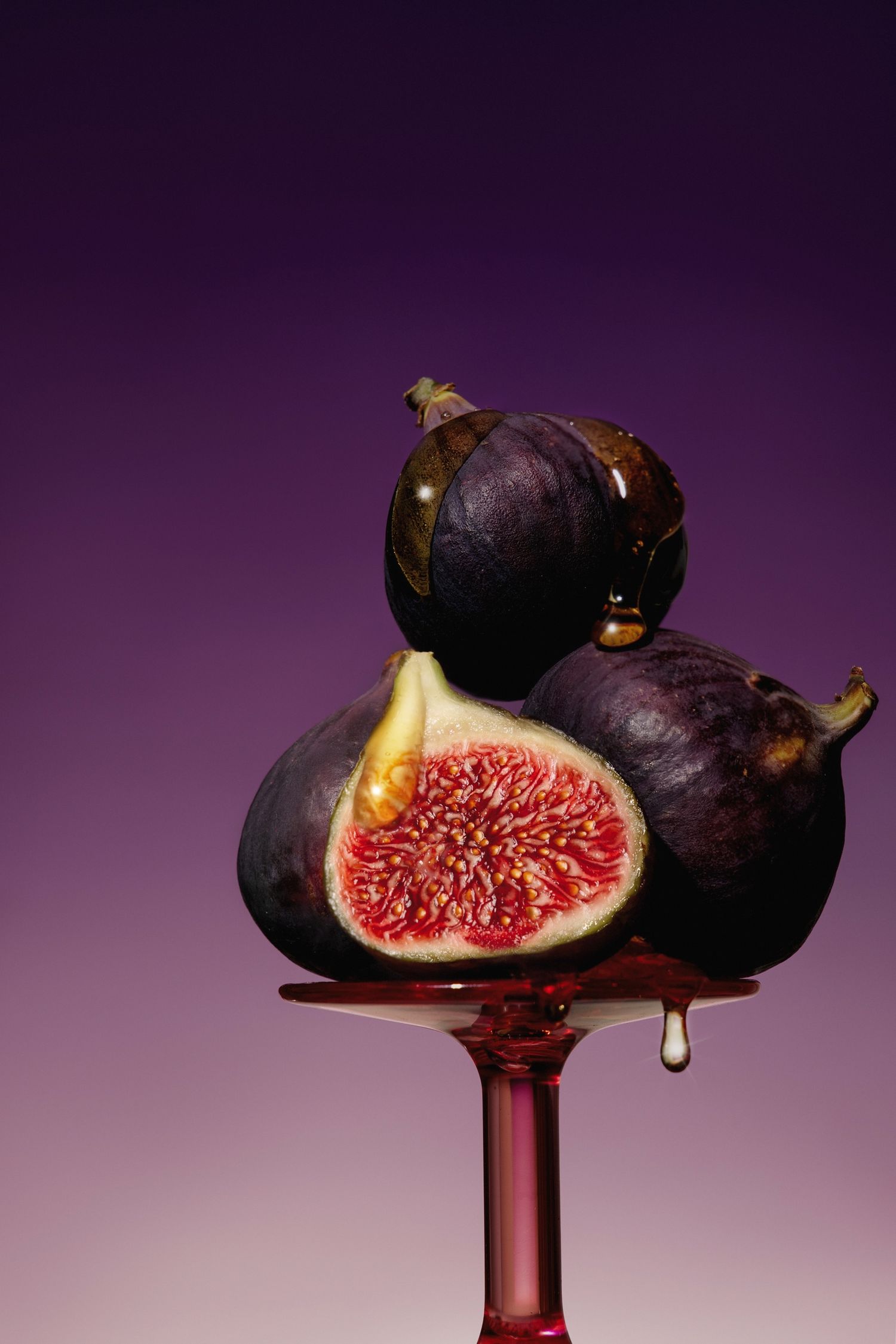 Fresh figs stacked on a wine glass, one sliced open revealing red interior, with honey dripping on purple background.