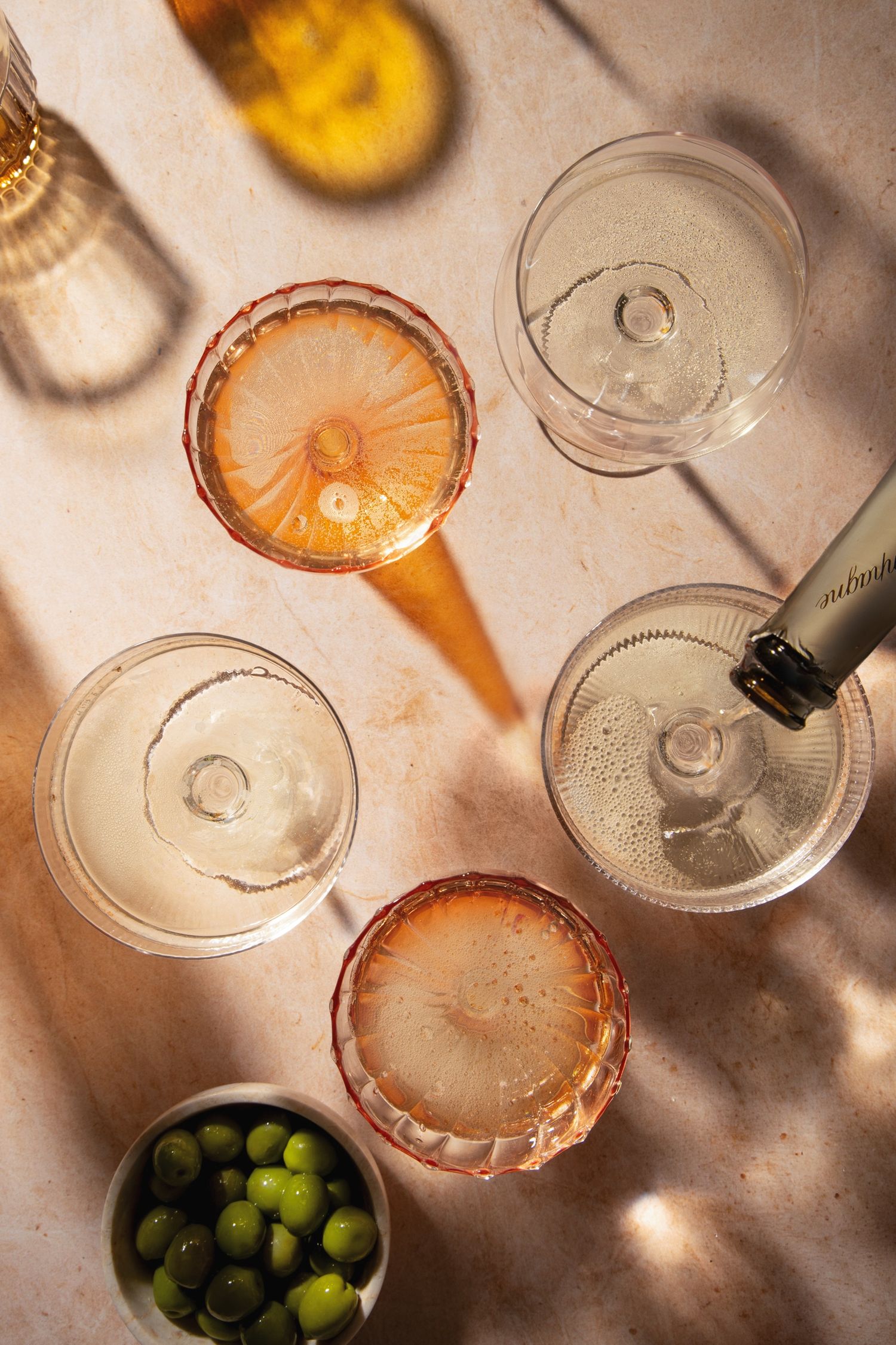 Overhead view of elegant cocktail glasses with champagne and rosé drinks, olives bowl, and golden shadows on marble surface.