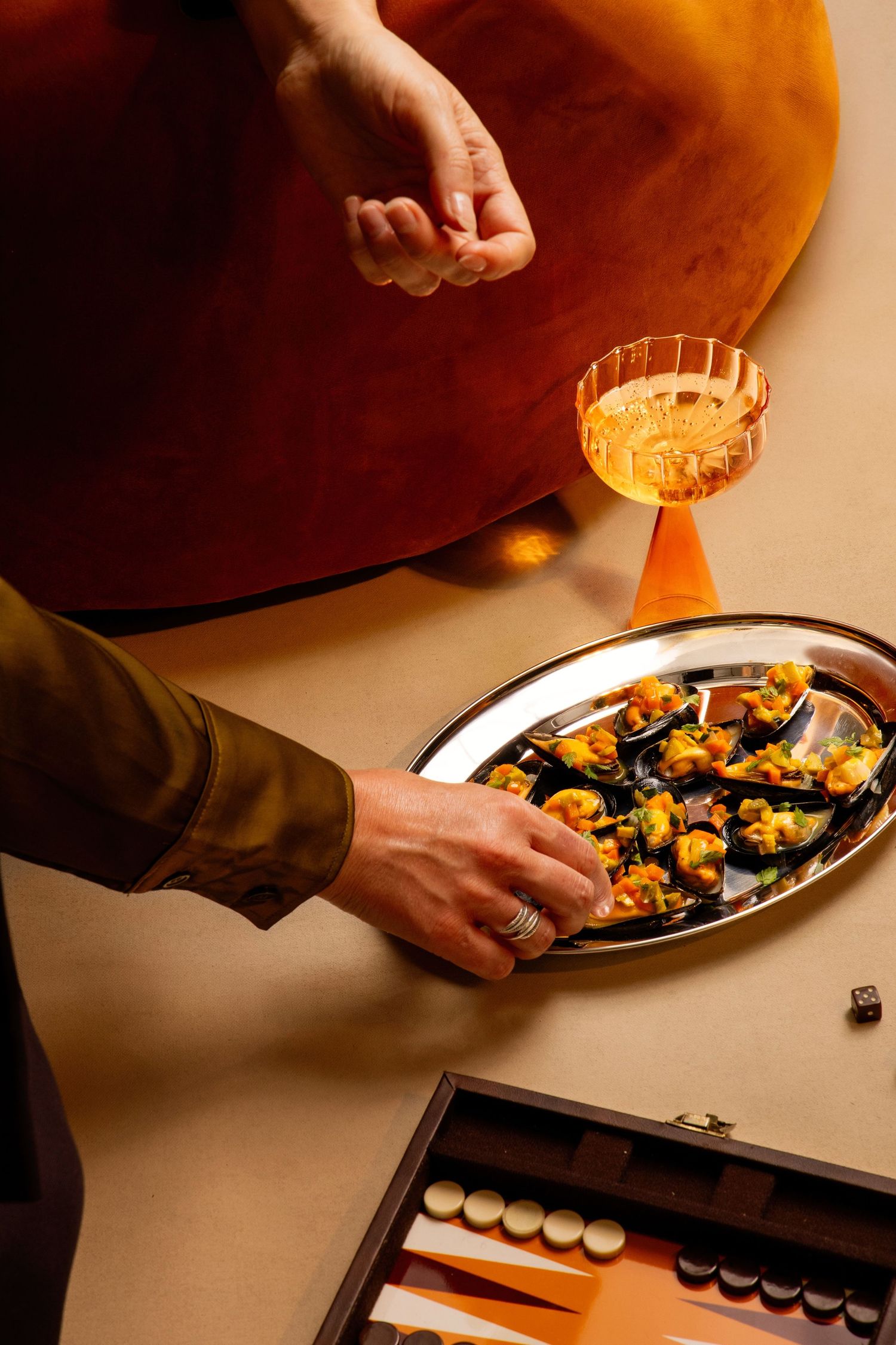Hands reaching for appetizers on a silver tray beside a cocktail glass and backgammon board on a beige table.